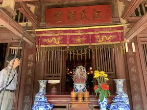 Bức ảnh chụp lối vào của một công trình kiến trúc cổ, dường như là một nhà thờ họ (từ đường) hoặc một ngôi đền. Toàn bộ công trình được làm bằng gỗ màu nâu sẫm. Ở trung tâm là một ban thờ được bài trí trang nghiêm. Phía trên lối vào có một bức hoành phi sơn son thếp vàng ghi ba chữ Hán lớn: 江氏祠堂 (Giang Thị Từ Đường), có nghĩa là nhà thờ của dòng họ Giang. Hai bên cột gỗ là hai câu đối chữ Hán. Câu đối bên phải là 敬績同垂南北 (Kính Tích Đồng Thùy Nam Bắc) và câu đối bên trái là 精神長在子孫身 (Tinh Thần Trường Tại Tử Tôn Thân). Phía trên ban thờ có treo một tấm y môn (cửa võng) bằng vải đỏ, thêu hình rồng vàng lộng lẫy. Trên ban thờ có một bát hương gốm sứ men lam lớn cắm đầy chân hương, hai chiếc lọ lộc bình gốm sứ men lam đối xứng hai bên. Một trong hai lọ cắm hoa tươi, bao gồm hoa cúc vàng và hoa mẫu đơn đỏ. Ngoài ra còn có các đài thờ bằng gỗ. Ở phía bên trái của bức ảnh, một người đàn ông lớn tuổi đội mũ lưỡi trai đang đứng và ngước nhìn lên kiến trúc của công trình.。（日本語訳）。古い建築物の入り口を写した写真で、一族の祠（祠堂）か寺院のようです。建物全体が濃い茶色の木材で造られています。中央には厳かに飾られた祭壇があります。入り口の上には、金箔を施した朱塗りの扁額があり、「江氏祠堂」（Giang Thị Từ Đường）という3つの大きな漢字が書かれています。これは江（Giang）氏一族の祠堂を意味します。両側の木の柱には、漢字の対聯（ついれん）が二つあります。右側の対聯は「敬績同垂南北」（Kính Tích Đồng Thùy Nam Bắc）、左側の対聯は「精神長在子孫身」（Tinh Thần Trường Tại Tử Tôn Thân）です。祭壇の上には、豪華な金色の龍が刺繍された赤い布製の幕（y môn/cửa võng）が掛けられています。祭壇の上には、線香の燃え残りがたくさん刺さった大きな青白磁の香炉が一つと、両脇に対称的に置かれた青白磁の飾り壺が二つあります。二つの壺のうちの一つには、黄色い菊と赤い牡丹を含む生花が生けられています。その他にも木製の祭具台があります。写真の左側には、野球帽をかぶった年配の男性が一人立っており、建物の建築を見上げています。