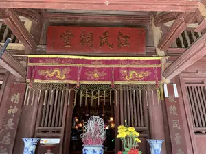 Bức ảnh chụp cận cảnh lối vào một công trình kiến trúc cổ của Việt Nam, có thể là một ngôi đình, chùa hoặc nhà thờ họ. Công trình được xây dựng chủ yếu bằng gỗ sơn màu đỏ sẫm. Phía trên cửa ra vào có một bức hoành phi lớn bằng gỗ sơn đỏ, khắc nổi ba chữ Hán mạ vàng: 江氏祠堂 (Giang Thị Từ Đường), nghĩa là nhà thờ của dòng họ Giang. Bên dưới hoành phi là một bức y môn bằng vải đỏ có thêu hình rồng vàng và các hoa văn trang trí. Hai bên cột nhà có treo hai câu đối bằng gỗ khắc chữ Hán. Ở phía trước lối vào, có một lư hương lớn bằng gốm sứ men lam trắng, bên trong cắm rất nhiều que hương đã được thắp. Bên cạnh lư hương là một bình hoa cúc vàng và hoa trang đỏ. Hai bên cửa là hai chiếc lọ lộc bình lớn bằng gốm sứ men lam trắng. Nhìn qua khe cửa có thể thấy không gian thờ tự ở bên trong với ánh đèn và các đồ vật trên ban thờ.。（日本語訳）。ベトナムの古い建築物の入り口を接写した写真で、村の集会所、寺院、または一族の祠堂である可能性があります。この建物は主に濃い赤色に塗られた木材で建てられています。出入り口の上には、赤く塗られた木製の大きな扁額があり、金メッキされた3つの漢字「江氏祠堂」（Giang Thị Từ Đường）が浮き彫りにされています。これは江（ザン）氏一族の祠堂を意味します。扁額の下には、金の龍と装飾文様が刺繍された赤い布製の幕があります。家の柱の両側には、漢字が刻まれた木製の対聯が2つ掛けられています。入り口の前には、青と白の染付磁器でできた大きな香炉があり、中には火が灯されたたくさんの線香が立てられています。香炉の隣には、黄色い菊と赤いサンタンカの花瓶があります。扉の両側には、青と白の染付磁器でできた大きな装飾用の壺が2つあります。扉の隙間からは、中の祭祀空間と、祭壇上の灯りや供物が見えます。