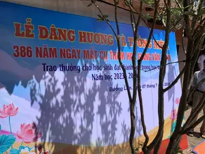 Bức ảnh chụp một tấm phông bạt lớn màu xanh được dựng ngoài trời, có thể là trong sân của một ngôi đình hoặc đền thờ tại làng cổ Đường Lâm, Sơn Tây. Tấm phông bạt này thông báo về một sự kiện đang diễn ra. Phía trước tấm phông là một cái cây, đổ bóng lên bề mặt. Nền sân được lát gạch. Phía bên phải có một người đàn ông đang đứng.。Nội dung chữ viết trên phông bạt:。LỄ DÂNG HƯƠNG VÀ TỔNG KẾT。386 NĂM NGÀY MẤT CỤ THÁM HOA GIANG VĂN MINH。Trao thưởng cho học sinh đạt thành tích cao trong học tập。Năm học 2023 - 2024。Đường Lâm, ngày 07 tháng 7 năm 2024。Hoạt động được mô tả là một buổi lễ kết hợp hai phần: lễ dâng hương tưởng niệm 386 năm ngày mất của danh nhân Thám hoa Giang Văn Minh và lễ trao thưởng cho học sinh có thành tích xuất sắc trong năm học 2023-2024. Các vật thể trong ảnh bao gồm phông bạt sự kiện, cây xanh, và hình ảnh trang trí hoa sen.。（日本語訳）。おそらく、ソンタイのドゥオンラム旧村にある村の集会所か祠の庭でしょう。この背景幕は、開催中のイベントについて告知しています。背景幕の前には木があり、その表面に影を落としています。庭の地面はレンガで舗装されています。右側には男性が一人立っています。背景幕に書かれた文字の内容：。献香式および総括式。タムホア・ザン・ヴァン・ミン公没後386年記念。学業で優秀な成績を収めた生徒への表彰。2023年～2024年度。ドゥオンラム、2024年7月7日。記述されている活動は、二部構成の式典です。すなわち、偉人タムホア・ザン・ヴァン・ミンの没後386年を記念する献香式と、2023-2024年度に優秀な成績を収めた生徒への表彰式です。写真に写っている物には、イベントの背景幕、緑の木、そして蓮の花の装飾画像が含まれます。