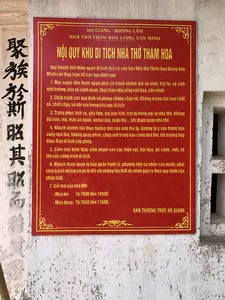 Bức ảnh chụp cận cảnh một tấm bảng nội quy màu đỏ với chữ vàng, được gắn trên một bức tường cũ kỹ. Đây là bảng nội quy dành cho khách tham quan tại Khu di tích Nhà thờ Thám hoa Giang Văn Minh, tọa lạc tại Đường Lâm. Bên cạnh tấm bảng là một hàng chữ Hán được viết dọc trên tường.。Nội dung văn bản trên bảng được ghi lại đầy đủ như sau:。Chữ Hán dọc: 聚族於斯貼其昭而。Bảng nội quy:。HỌ GIANG - ĐƯỜNG LÂM。NHÀ THỜ THÁM HOA GIANG VĂN MINH。NỘI QUY KHU DI TÍCH NHÀ THỜ THÁM HOA。Quý khách đến thăm quan di tích lịch sử-văn hóa Nhà thờ Thám hoa Giang Văn Minh cần thực hiện tốt các quy định sau:。1. Mọi người đến tham quan phải có trách nhiệm giữ an ninh, trật tự, vệ sinh môi trường, bảo vệ cảnh quan, thực hiện nếp sống văn hóa, văn minh.。2. Chấp hành các quy định về phòng chống cháy nổ. Không mang các loại chất nổ, chất cháy, vũ khí vào trong khu vực di tích.。3. Trang phục lịch sự, phù hợp, giữ yên tĩnh, trật tự trong nội tự nhà thờ. Không đội nón, mũ, mặc áo ngắn, áo hai dây, quần đùi... khi tham quan nhà thờ.。4. Khách chiêm bái theo hướng dẫn của ông thủ từ. Không tự ý vào trong hậu cung nhà thờ, không quay phim, chụp ảnh trong hậu cung nhà thờ, trừ trường hợp được Ban di tích cho phép.。5. Cấm mọi hình thức xâm phạm vào các hiện vật, hái hoa, bẻ cành, viết, vẽ lên các công trình di tích.。6. Khách tham quan tự bảo quản hành lý, phương tiện cá nhân của mình; phải chịu trách nhiệm pháp lý đối với những tổn thất do mình gây ra theo quy định của pháp luật.。7. Giờ mở cửa nhà thờ:。- Mùa hè: Từ 7h00 đến 18h00。- Mùa đông: Từ 7h30 đến 17h00.。BAN THƯỜNG TRỰC HỌ GIANG。（日本語訳）。金色の文字が書かれた赤い規則掲示板のクローズアップ写真で、古びた壁に取り付けられています。これは、ドゥオンラムに位置するタムホア・ザン・ヴァン・ミン祠堂遺跡地区の拝観者向けの規則掲示板です。掲示板の隣には、壁に縦書きされた一行の漢文があります。掲示板の文章内容は、以下の通りです。縦書きの漢文：聚族於斯貼其昭而。規則掲示板：。ザン氏族 - ドゥオンラム。タムホア・ザン・ヴァン・ミン祠堂。タムホア祠堂遺跡地区 規則。タムホア・ザン・ヴァン・ミン祠堂歴史文化遺跡を拝観される皆様は、以下の規定を遵守してください。1. 拝観に来られたすべての方は、安全、秩序、環境衛生を保ち、景観を保護し、文化的で文明的な生活様式を実践する責任を負わなければなりません。2. 火災・爆発防止に関する規定を遵守すること。爆発物、可燃物、武器を遺跡区域内に持ち込まないこと。3. 礼儀正しく、適切な服装をし、祠堂内では静粛にし、秩序を保つこと。祠堂を拝観する際は、ノン（笠）、帽子をかぶったり、短いシャツ、キャミソール、ショートパンツなどを着用したりしないこと。4. 参拝客は祠守の案内に従うこと。遺跡管理委員会から許可された場合を除き、祠堂の後宮に勝手に入ったり、後宮内でビデオ撮影や写真撮影をしたりしないこと。5. 展示物へのあらゆる形式の侵害、花を摘むこと、枝を折ること、遺跡の建造物への落書きを禁じます。6. 拝観者は自身の荷物、個人の乗り物を自己管理すること。自身が引き起こした損害に対しては、法律の規定に従い、法的責任を負わなければなりません。7. 祠堂の開門時間：。- 夏季：7時00分から18時00分まで。- 冬季：7時30分から17時00分まで。ザン氏族常務委員会