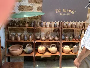 Trên tấm biển hiệu màu nâu có ghi dòng chữ nghệ thuật màu vàng: 'Dệt hương' và 'Di sản Việt'. Bối cảnh là một gian hàng trưng bày các sản phẩm gốm sứ thủ công, được đặt trên những chiếc kệ gỗ nhiều tầng. Phía sau kệ là một bức tường đá ong cổ kính. Các sản phẩm bao gồm rất nhiều bình, lọ gốm với nhiều hình dáng và màu sắc nâu khác nhau ở các tầng trên. Tầng kệ dưới cùng trưng bày các ấm trà bằng đất nung và một chiếc nồi đất lớn. Một chiếc lá chuối lớn được dùng để trang trí ở góc trên bên trái. Ở phía bên phải, một người đàn ông đang đứng xem các sản phẩm.。（日本語訳）。茶色の看板には、「Dệt hương」と「Di sản Việt」という黄色い芸術的な文字が書かれています。背景は、手作りの陶磁器製品を展示するブースで、それらは何段にもなった木製の棚の上に置かれています。棚の後ろには、古風なラテライトの壁があります。製品には、上の段にある様々な形と異なる茶色の色合いを持つ多くの陶器の壺や瓶が含まれています。一番下の棚には、素焼きの急須と大きな土鍋が展示されています。左上の角には、大きなバナナの葉が装飾として使われています。右側では、一人の男性が立って製品を見ています。