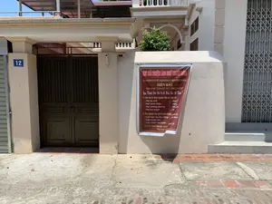 Bức ảnh chụp phía trước một tòa nhà, có thể là một cơ sở kinh doanh, với biển số nhà màu xanh ghi số '12' gắn trên cột cổng. Trên bức tường màu kem, có treo một tấm bạt lớn màu nâu đỏ quảng cáo dịch vụ.。Nội dung trên tấm bạt được ghi lại như sau:。- Tiêu đề: 'KHU TRẢI NGHIỆM LÀNG NGHỀ TRUYỀN THỐNG'。- Tên cơ sở: 'CƠ SỞ SẢN XUẤT BÁNH KẸO TRUYỀN THỐNG HIỀN BAO'。- Điện thoại: 0359.037.107 - 0989.915.081。Tấm bạt mời khách hàng đến trải nghiệm các hoạt động:。1. Làm kẹo tự chọn nguyên liệu từ các loại hạt: Hạt óc chó, hạt hạnh nhân, hạt hướng dương, hạt lúa mạch (một số loại hạt khác bị che khuất).。2. Trải nghiệm các nghề truyền thống: Làm kẹo lạc thủ công, làm bánh sắn, làm bánh rán nước, bánh tẻ, chè lam (một số mục bị che khuất). Có ghi chú 'MANG VỀ'.。Phần dưới cùng của tấm bạt có ghi 'CHI PHÍ THỎA THUẬN'.。Cảnh vật xung quanh bao gồm một cổng sắt lớn màu nâu sẫm đang khóa, một cửa cuốn xếp bằng kim loại ở bên phải, và vỉa hè bằng bê tông và gạch. Không có người hay hoạt động nào đang diễn ra trong ảnh.。（日本語訳）。写真は、ある建物の正面を撮影したもので、おそらく事業所であり、門柱には「12」と書かれた青い住居表示板が取り付けられています。クリーム色の壁には、サービスを宣伝する赤褐色の大きな垂れ幕が掛けられています。垂れ幕に記載されている内容は以下の通りです。- 見出し：「伝統工芸村体験エリア」。- 事業所名：「ヒエン・バオ伝統菓子製造所」。- 電話番号：0359.037.107 - 0989.915.081。垂れ幕は、以下の活動を体験するよう顧客を誘っています。1. 各種のナッツから材料を自分で選んで飴作り：クルミ、アーモンド、ヒマワリの種、大麦（他のいくつかのナッツは隠れて見えません）。2. 伝統的な職業の体験：手作りピーナッツキャンディー作り、キャッサバ餅作り、バイン・ラン・ヌオック（水揚げドーナツ）作り、バイン・テー、チェー・ラム（いくつかの項目は隠れて見えません）。「持ち帰り」という注記があります。垂れ幕の最下部には「費用は応相談」と書かれています。周囲の光景には、施錠された濃い茶色の大きな鉄の門、右側にある金属製の折りたたみ式シャッター、そしてコンクリートとレンガでできた歩道が含まれています。写真の中では、人や活動は行われていません。