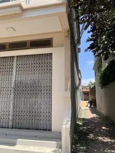 Bối cảnh là một con ngõ nhỏ, hẹp, được lát gạch, nằm giữa hai dãy nhà. Ngôi nhà bên trái là một công trình hiện đại, sơn màu kem, có cửa sắt kéo màu xám đang đóng. Bên phải là bức tường của một ngôi nhà khác và một cây lớn đổ bóng xuống lối đi. Ở phía xa trong con ngõ, một người đội nón lá đang làm việc, có vẻ như đang cào và phơi nông sản (như thóc hoặc đậu) trên một tấm bạt trắng trải trên mặt đất. Phía cuối ngõ có thể nhìn thấy những ngôi nhà khác với mái ngói đỏ và một bồn nước. Trên tường có một biển số nhà nhỏ màu xanh ghi số '4'.。（日本語訳）。背景は、二つの家並みの間にある、レンガが敷かれた小さく狭い路地です。左側の家は現代的な建物で、クリーム色に塗られ、灰色の引き戸の鉄の扉が閉まっています。右側は別の家の壁と、通路に影を落とす大きな木です。路地の奥では、ノンラーをかぶった人が作業をしており、地面に広げられた白いシートの上で農産物（籾や豆など）を熊手でかき集めて干しているようです。路地の突き当りには、赤い瓦屋根の他の家々と貯水タンクが見えます。壁には、「4」と書かれた小さな青い家屋番号プレートがあります。