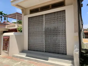 Bức ảnh chụp mặt tiền của một ngôi nhà vào ban ngày, có vẻ là một cơ sở sản xuất. Ngôi nhà được sơn màu kem, có một bộ cửa sắt kéo lớn màu xám đang đóng kín. Phía trước cửa là các bậc tam cấp bằng xi măng. Ở bên trái lối vào, trên một cột trụ, có treo một tấm biển thông báo màu đỏ và trắng. Nội dung trên biển hiệu được ghi lại như sau:。CƠ SỞ SẢN XUẤT BÁNH KẸO TRUYỀN THỐNG。HIỀN BẢO。Điện thoại: 0359.037.107 - 0989.915.081。Quý khách đến Ba Vì ghé Cơ sở Hiền Bảo đi tham quan。1. Làm kẹo tự chọn nguyên liệu các loại hạt:。- Hạt óc chó - Hạt điều。- Hạt hạnh nhân - Hạt bí。- Hạt hướng dương - Hạt vừng。- Hạt lúa mạch - Hạt gạo。2. Trải nghiệm các nghề truyền thống:。- Làm kẹo lạc thủ công - Làm b... (chữ bị mờ)。- Làm bánh sắn - Làm bánh rán nước。- Làm ch... (chữ bị mờ)。CHI PHÍ THỎA THUẬN - CÓ SẢN PHẨM MANG VỀ。Xin trân trọng。Bối cảnh xung quanh cho thấy các ngôi nhà khác có mái ngói, ban công, và cây cối xanh tươi bao gồm cả cây cau, dưới bầu trời xanh. Ở phía bên phải của ngôi nhà, trong một khoảng sân nhỏ, có nông sản (có thể là các loại hạt được liệt kê trên biển hiệu) đang được phơi khô trên một tấm bạt trắng trải trên mặt đất. Hoạt động chính có thể nhìn thấy là việc phơi nông sản.。（日本語訳）。日中に撮影されたある家の正面の写真で、生産施設のようです。家はクリーム色に塗られており、大きな灰色の鉄製シャッターが閉まっています。ドアの前にはセメント製の階段があります。入り口の左側、柱の上には、赤と白の看板が掲げられています。看板の内容は以下の通りです。伝統菓子製造所。ヒエン・バオ。電話番号：0359.037.107 - 0989.915.081。お客様がバヴィにお越しの際は、ヒエン・バオ製造所にお立ち寄りいただき、ご見学ください。1. 各種ナッツ類を材料に選んで作るお菓子作り：。- クルミ - カシューナッツ。- アーモンド - カボチャの種。- ヒマワリの種 - ゴマ。- 大麦 - 米粒。2. 伝統的な職業体験：。- 手作りピーナッツ菓子 - b...作り（文字が不鮮明）。- キャッサバ餅作り - 水揚げドーナツ作り。- ch...作り（文字が不鮮明）。費用は応相談 - お持ち帰り製品あり。敬具。周囲の背景には、青空の下、瓦屋根やバルコニーのある他の家々、そしてビンロウを含む青々とした木々が見えます。家の右側、小さな庭では、農産物（看板に記載されているナッツ類かもしれません）が地面に敷かれた白いシートの上で天日干しされています。見て取れる主な活動は、農産物の天日干しです。