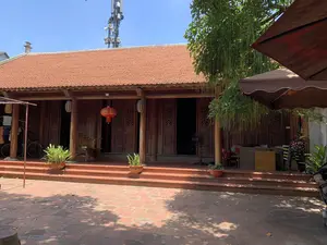 Bức ảnh chụp một ngôi nhà gỗ truyền thống của Việt Nam vào một ngày nắng. Ngôi nhà có mái lợp ngói vảy cá màu đỏ đặc trưng và kết cấu hoàn toàn bằng gỗ, từ cột nhà đến các bộ cửa được chạm khắc hoa văn. Phía trước nhà là một khoảng sân rộng được lát gạch đỏ. Trên hiên nhà có bậc tam cấp, được trang trí bằng vài chậu cây cảnh. Một chiếc đèn lồng màu đỏ và một chiếc đèn lồng khác được treo trước cửa. Bên trong sân, có một chiếc ô lớn màu nâu che nắng và bóng cây râm mát. Các vật dụng sinh hoạt như xe đạp, giá phơi quần áo, và một chiếc quạt điện cho thấy đây là nơi ở hoặc kinh doanh. Ở phía sau ngôi nhà, một cột tháp viễn thông cao với các thiết bị và loa phát thanh nổi bật trên nền trời xanh. Khung cảnh yên tĩnh, thể hiện sự kết hợp giữa kiến trúc cổ truyền và các yếu tố hiện đại.。（日本語訳）。ある晴れた日に撮影された、ベトナムの伝統的な木造家屋の写真。この家は、特徴的な赤い魚の鱗瓦の屋根を持ち、柱から模様が彫られた扉に至るまで、完全に木で造られている。家の前には、赤いレンガが敷かれた広い庭がある。軒先には三段の階段があり、いくつかの鉢植えの観葉植物で飾られている。赤い提灯と、もう一つの提灯が戸口の前に吊るされている。庭の中には、日差しを遮る大きな茶色の傘と、涼しい木陰がある。自転車、物干し台、扇風機といった生活用品は、ここが住居または商業施設であることを示している。家の後ろには、青空を背景に、機器や拡声器を備えた高い通信塔がそびえ立っている。静かな光景は、伝統建築と現代的な要素の融合を表している。