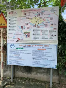 Đây là hình ảnh chụp một tấm biển thông tin du lịch lớn dành cho du khách tại Làng cổ Đường Lâm, Sơn Tây, Hà Nội. Tấm biển được đặt ngoài trời, bên cạnh một bức tường đá và có cây cối xung quanh. Một phần của biển báo giao thông hình tam giác màu vàng viền đỏ có chữ "CHẬM" và một chiếc gương cầu lồi cũng xuất hiện trong ảnh.。Tấm biển bao gồm hai phần chính: một bản đồ du lịch và một bảng quy tắc ứng xử.。**Phần 1: Bản đồ du lịch "Welcome to Làng cổ Đường Lâm"**。Bản đồ này cung cấp thông tin chi tiết về các địa điểm tham quan, nhà hàng, nhà cổ và các hoạt động trải nghiệm trong làng, được chia thành các thôn: Đông Sàng, Đoài Giáp, Mông Phụ, Cam Thịnh, và Cam Lâm. Các địa điểm được liệt kê bao gồm:。*   **Nhà cổ (Traditional houses):**。1.  Nhà cổ ông Nguyễn Văn Hùng。2.  Nhà cổ ông Hà Hữu Thể。3.  Nhà cổ ông Hà Nguyên Huyến。4.  Nhà cổ ông Hà Văn Vĩnh。5.  Nhà cổ bà Dương Thị Lan。6.  Nhà cổ ông Cao Văn Toàn。7.  Nhà cổ ông Giang Văn Thuận。8.  Nhà cổ ông Kiều Anh Ban。9.  Nhà cổ bà Vũ Thị Âm。10. Nhà cổ ông Nguyễn Huy Chương。*   **Homestay:**。24. Nok Homestay。25. Duong Lam Village。26. Xomhe Villa。27. Ca phe Lang Homestay。*   **Nhà hàng - Cà phê (Restaurant - Cafe):**。11. Nhà ông Phan Văn Hài。12. Công ty du lịch Đường Lâm。13. Nhà bà Hà Thị Điền。14. Nhà bà Kiều Thị Tính。15. Nhà bà Khanh。16. Bếp làng Đường Lâm。17. Cà phê ĐOÀI。18. Cà phê Làng。*   **Trải nghiệm & học tập ngành nghề địa phương (Local industry study & experience):**。19. Nhà ông Hiền Bao (làm bánh kẹo)。20. Nhà ông Giang Vĩnh Phúc (làm chè lam, chè kho, bánh tẻ, bánh gai)。21. Nhà ông Phan Văn Thách (làm tương)。22. Điểm giới thiệu bán sản phẩm OCOP。23. Phát Studio, Không gian sáng tạo ĐOÀI (Art Work Shop, Art gallery)。*   **Các địa điểm khác:** Cổng làng Đông Sàng, Chùa Mía, Đình Tổng, Đền Phùng Hưng, Nhà thờ Mông Phụ, Cổng Mông Phụ, Lăng Ngô Quyền, v.v.。Bản đồ cũng đề xuất hai tuyến đường tham quan (route A, route B).。**Phần 2: Bảng "QUY TẮC ỨNG XỬ NƠI CÔNG CỘNG TRÊN ĐỊA BÀN THÀNH PHỐ HÀ NỘI"**。Bảng này trích dẫn các quy định từ Quyết định số 1665/QĐ-UBND ngày 10/3/2017 của UBND thành phố Hà Nội, chia thành các mục "NÊN LÀM" và "KHÔNG NÊN LÀM".。*   **ĐIỀU 3. QUY TẮC ỨNG XỬ CHUNG:**。*   **NÊN LÀM:** Tuân thủ pháp luật, nội quy; Tôn trọng không gian chung; Ứng xử lịch thiệp, thân thiện; Trang phục lịch sự; Quan tâm, giúp đỡ người khuyết tật, người già, trẻ em; Đấu tranh, bảo vệ lẽ phải; Tôn trọng, bảo vệ cảnh quan môi trường.。*   **KHÔNG NÊN LÀM:** Vi phạm nội quy; Gây ồn ào; Kích động, đe dọa, bạo lực; Nói tục, chửi bậy; Hút thuốc, khạc nhổ; Xả rác bừa bãi; Phá hoại cây xanh, cảnh quan; Tự tiện sử dụng không gian công cộng; Viết, vẽ bậy; Thả rông vật nuôi; Sử dụng vũ khí, chất gây nổ trái phép.。*   **ĐIỀU 6. TẠI CƠ SỞ TÍN NGƯỠNG, TÔN GIÁO:**。*   **NÊN LÀM:** Tôn trọng tự do tín ngưỡng; Giữ gìn giá trị truyền thống; Chấp hành quy định tại nơi thờ tự; Đi nhẹ, nói khẽ, giữ trật tự.。*   **KHÔNG NÊN LÀM:** Thực hành mê tín dị đoan; Lợi dụng tín ngưỡng để trục lợi; Xâm hại cảnh quan, không gian tín ngưỡng; Mặc trang phục hở hang, không phù hợp.。*   **ĐIỀU 11. TẠI KHU VUI CHƠI, GIẢI TRÍ, ĐIỂM THAM QUAN DU LỊCH:**。*   **NÊN LÀM:** Mặc trang phục phù hợp; Thể hiện tình cảm đúng mực; Tuân thủ hướng dẫn; Mua, bán hàng đúng nơi quy định; Bảo vệ cảnh quan môi trường; Lấy sách hướng dẫn khi cần thiết; Tôn trọng văn hóa, phong tục địa phương.。*   **KHÔNG NÊN LÀM:** Chen lấn, xô đẩy; Ăn uống, ngủ nghỉ tùy tiện; Tổ chức hoạt động trái quy định; Sử dụng đồ chơi bạo lực, nguy hiểm; Tranh giành, chèo kéo khách; Nâng giá hàng hóa, dịch vụ.。（日本語訳）。これは、ハノイ市ソンタイ、ドゥオンラム古村にある観光客向けの大きな観光案内板を撮影した写真です。案内板は屋外の石垣の隣に設置されており、周りには木々があります。案内板は、観光地図と行動規範の2つの主要な部分で構成されています。**第1部：観光地図「ドゥオンラム古村へようこそ」**。この地図は、村内の観光スポット、レストラン、古民家、体験活動に関する詳細な情報を提供しており、ドンサン、ドアイジャップ、モンフー、カムティン、カムラムの各集落に分かれています。記載されている場所は以下の通りです。*   **古民家 (Traditional houses):**。1.  グエン・ヴァン・フン氏の古民家。2.  ハ・フー・テー氏の古民家。3.  ハ・グエン・フエン氏の古民家。4.  ハ・ヴァン・ヴィン氏の古民家。5.  ズオン・ティ・ラン氏の古民家。6.  カオ・ヴァン・トアン氏の古民家。7.  ジャン・ヴァン・トゥアン氏の古民家。8.  キエウ・アイン・バン氏の古民家。9.  ヴー・ティ・アム氏の古民家。10. グエン・フイ・チュオン氏の古民家。*   **ホームステイ:**。24. ノック・ホームステイ。25. ドゥオンラム・ビレッジ。26. ソムヘ・ヴィラ。27. カフェ・ラン・ホームステイ。*   **レストラン - カフェ (Restaurant - Cafe):**。11. ファン・ヴァン・ハイ氏の家。12. ドゥオンラム観光会社。13. ハ・ティ・ディエン氏の家。14. キエウ・ティ・ティン氏の家。15. カイン氏の家。16. ドゥオンラム村の台所。17. ドアイ・カフェ。18. 村カフェ。*   **地元の産業の学習と体験 (Local industry study & experience):**。19. ヒエン・バオ氏の家（菓子作り）。20. ジャン・ヴィン・フック氏の家（チェーラム、チェーコー、バインテー、バインガイ作り）。21. ファン・ヴァン・タック氏の家（トゥオン（味噌）作り）。22. OCOP製品の紹介・販売所。23. ファットスタジオ、ドアイクリエイティブスペース（アートワークショップ、アートギャラリー）。*   **その他の場所:** ドンサン村の門、ミア寺、トン村の亭、フン・フン祠、モンフー教会、モンフー門、ゴ・クエン陵、など。地図はまた、2つの観光ルート（ルートA、ルートB）を提案しています。**第2部：「ハノイ市内の公共の場所における行動規範」**。この表は、ハノイ市人民委員会による2017年3月10日付の決定第1665/QĐ-UBND号からの規定を引用しており、「すべきこと」と「すべきでないこと」の項目に分かれています。*   **第3条. 一般的な行動規範:**。*   **すべきこと:** 法律、規則を遵守する。公共の空間を尊重する。礼儀正しく、親しみやすく振る舞う。品位のある服装をする。障害者、高齢者、子供に関心を持ち、助ける。正義のために闘い、守る。景観環境を尊重し、保護する。*   **すべきでないこと:** 規則に違反する。騒音を立てる。扇動、脅迫、暴力行為。下品な言葉、罵詈雑言を言う。喫煙、痰を吐く。ゴミをポイ捨てする。樹木、景観を破壊する。公共の空間を勝手に使用する。落書きをする。ペットを放し飼いにする。武器、爆発物を不法に使用する。*   **第6条. 信仰、宗教施設にて:**。*   **すべきこと:** 信仰の自由を尊重する。伝統的価値を維持する。礼拝所の規定を遵守する。静かに歩き、静かに話し、秩序を保つ。*   **すべきでないこと:** 迷信的な行為を行う。信仰を利用して私利を貪る。景観、信仰の空間を侵害する。露出の多い、不適切な服装をする。*   **第11条. 遊園地、娯楽施設、観光地にて:**。*   **すべきこと:** 適切な服装をする。節度ある愛情表現をする。指示に従う。規定の場所で商品を売買する。景観環境を保護する。必要に応じて案内書を取る。地元の文化、風習を尊重する。*   **すべきでないこと:** 割り込み、押し合いをする。勝手に飲食、休息、睡眠をとる。規定に反する活動を組織する。暴力的な、危険な玩具を使用する。客の奪い合い、客引きをする。商品、サービスの価格を吊り上げる。