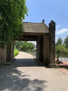 Bức ảnh chụp một chiếc cổng thành cổ, được xây dựng bằng đá và gỗ với kiến trúc truyền thống. Mái cổng lợp ngói âm dương đã nhuốm màu thời gian. Cánh cổng gỗ lớn đang mở, để lộ ra con đường bê tông dẫn vào bên trong, hai bên đường là hàng cây xanh mát. Phía xa trên con đường có một biển báo giao thông màu vàng cảnh báo khúc cua. Bên phải cổng là một ao nước, xa hơn là những hàng cây dừa. Gần đó có một tấm bia ghi thông tin đặt trên bệ nhưng không thể đọc được chữ. Bên trái là một tán cây lớn rậm rạp. Bầu trời trong xanh, quang đãng, cho thấy đây là một ngày nắng đẹp. Khung cảnh vắng lặng, mang vẻ cổ kính, yên bình.。（日本語訳）。写真は、伝統的な建築様式で石と木で建てられた古い城門を写している。門の屋根は陰陽瓦で葺かれており、時の流れを感じさせる色合いになっている。大きな木製の門扉は開いており、中へと続くコンクリートの道が現れている。道の両側には涼しげな緑の並木がある。道の先の方には、カーブを警告する黄色い交通標識がある。門の右側には池があり、その向こうにはココヤシの木々が並んでいる。その近くには台座の上に情報が刻まれた石碑があるが、文字を読むことはできない。左側には、葉が鬱蒼と茂った大きな木がある。空は青く澄み渡っており、晴れた美しい日であることがわかる。辺りは静まり返り、古風で平和な趣を醸し出している。