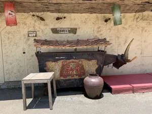 Bối cảnh là một không gian trưng bày ngoài trời, có lẽ là trước một cửa hàng hoặc xưởng thủ công mỹ nghệ ở làng cổ Đường Lâm. Bức tường có màu vàng, làm bằng vật liệu trông giống như đất trộn rơm, và mái hiên được lợp bằng tre, nứa đan. Ở trung tâm của bức ảnh là một chiếc xe đẩy trang trí độc đáo, có thể dùng làm quầy hàng. Xe có phần thân bằng kim loại hoặc gỗ sẫm màu, được trang trí bằng các họa tiết màu đỏ và vàng đã cũ, gợi nhớ đến hoa văn trên trống đồng Đông Sơn. Mái che của xe được lợp bằng ngói âm dương. Bên cạnh xe là một chiếc bàn gỗ nhỏ, mộc mạc và một chiếc chum sành lớn màu nâu. Treo trên mái hiên có hai chiếc đèn lồng (một đỏ, một xanh) và trên tường có gắn các vật trang trí hình con kiến bằng kim loại. Không có người hay hoạt động nào đang diễn ra, đây là một cảnh tĩnh. Các chữ viết có thể nhận dạng bao gồm: biển gỗ "DUONG LAM GIFT", biển gỗ lớn hơn ghi "DUONG LAM LACQUER ART" và hàng chữ Nhật "ドラム芸 술" (Nghệ thuật Trống), và chữ "DUONG LAM VILLAGE" trên chiếc đèn lồng màu đỏ.。（日本語訳）。背景は屋外の展示スペースで、おそらくドゥオンラム旧村にある手工芸品店または工房の前です。壁は黄色で、藁を混ぜた土のような素材でできており、軒は竹や細竹を編んで葺かれています。写真の中央には、屋台として使用できるユニークな装飾が施された手押し車があります。車体は金属または濃い色の木でできており、ドンソン銅鼓の文様を彷彿とさせる、古びた赤と黄色の模様で装飾されています。車の屋根は陰陽瓦で葺かれています。車の隣には、素朴な小さな木製のテーブルと、大きな茶色の陶器の壺があります。軒には2つの提灯（1つは赤、1つは緑）が吊るされ、壁には金属製のアリの形の装飾品が取り付けられています。人や活動は見られず、静的な光景です。認識できる文字には、木製の看板「DUONG LAM GIFT」、より大きな木製の看板「DUONG LAM LACQUER ART」、日本の文字「ドラム芸 술」（太鼓の芸術）、そして赤い提灯の文字「DUONG LAM VILLAGE」が含まれます。