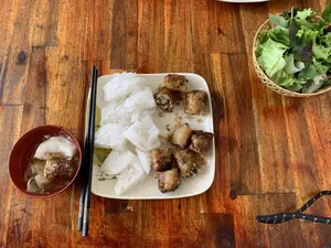 Hình ảnh chụp một bữa ăn được bày trên bàn gỗ. Món ăn chính là bún nem rán, một món ăn của Việt Nam. Trên một chiếc đĩa hình chữ nhật màu trắng ngà có hoa văn rạn, có bún tươi và nem rán đã được cắt thành từng miếng. Bên trái là một bát nước chấm màu đỏ, bên trong có một miếng nem. Một đôi đũa màu đen được đặt cạnh đĩa thức ăn. Ở phía trên bên phải là một chiếc rổ tre đựng các loại rau sống ăn kèm, bao gồm xà lách và tía tô. Khung cảnh cho thấy một bữa ăn đang được thưởng thức.。（日本語訳）。木製のテーブルの上に並べられた食事の写真。メイン料理はベトナム料理の一つであるブンネムザンです。ひび割れ模様のある象牙色の長方形の皿には、生のブンと、一切れずつに切られた揚げ春巻きがあります。左側には赤いタレの入ったお椀があり、その中に春巻きが一切れ入っています。料理の皿の隣に黒い箸が置かれています。右上には、レタスやシソなどの付け合わせの生野菜が入った竹かごがあります。食事が楽しまれている様子がうかがえる光景です。