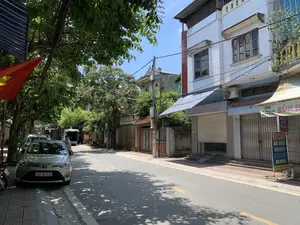 Bối cảnh là một con phố tại Việt Nam vào một ngày nắng. Dựa vào các biển hiệu, đây là Phố Trần Hưng Đạo, có khả năng thuộc thị xã Sơn Tây. Con phố có nhiều nhà ống cao tầng san sát, hai bên đường là hàng cây xanh tỏa bóng mát xuống vỉa hè và lòng đường. Khung cảnh khá yên tĩnh, không có hoạt động mua bán tấp nập. Một số cửa hàng, như quán ăn ở số 36, đang đóng cửa bằng cửa cuốn và cửa xếp.。Các vật thể và hoạt động trong ảnh bao gồm:。- **Loại địa điểm:** Một con phố đô thị với nhà ở và cửa hàng kinh doanh.。- **Hoạt động:** Một chiếc ô tô màu bạc đang đỗ bên lề đường. Một người đàn ông đang đi bộ ở phía xa.。- **Vật thể:** Nhà ống, cây xanh, cột điện với dây điện chằng chịt, một lá cờ Tổ quốc Việt Nam (cờ đỏ sao vàng), ô tô, xe máy, và các biển hiệu.。Nội dung các văn bản có thể đọc được:。- Biển chỉ dẫn ngõ: 'NGÕ 38 PHỐ TRẦN HƯNG ĐẠO'.。- Biển hiệu quán ăn tại số 36: 'BÚN BÒ HUẾ', 'BÁNH BỘT LỌC', 'LƯƠN RIM BIỂN', 'MỰC RIM ME', 'CHẢ CUA HUẾ'. Địa chỉ được ghi là '36 TRẦN HƯNG ĐẠO' cùng số điện thoại 'ĐT: 039 756 166 43'.。- Biển hiệu khác: 'CHÁO GẦU' với số điện thoại '035 9112016'.。（日本語訳）。背景は、ある晴れた日のベトナムの通りです。看板によると、ここはチャン・フン・ダオ通りで、ソンタイ市社に属している可能性があります。通りには高層のチューブハウスが密集しており、道の両側には街路樹が歩道と車道に涼しい木陰を落としています。風景はかなり静かで、賑やかな売買活動はありません。36番地の食堂など、いくつかの店はシャッターや折りたたみ式のドアで閉まっています。写真の中の物体と活動には以下が含まれます：。- **場所の種類：** 住居と商業店舗がある都市の通り。- **活動：** 銀色の車が道端に駐車しています。遠くで男性が一人歩いています。- **物体：** チューブハウス、緑の木々、複雑に絡み合った電線のある電柱、ベトナム国旗（金星紅旗）、自動車、バイク、そして看板。読み取れるテキストの内容：。- 路地の案内標識：「NGÕ 38 PHỐ TRẦN HƯNG ĐẠO」（チャン・フン・ダオ通り38番路地）。- 36番地の食堂の看板：「BÚN BÒ HUẾ」（ブンボーフエ）、「BÁNH BỘT LỌC」（バインボットロック）、「LƯƠN RIM BIỂN」（ウナギの煮付け）、「MỰC RIM ME」（イカのタマリンド煮）、「CHẢ CUA HUẾ」（フエ風カニすり身揚げ）。住所は「36 TRẦN HƯNG ĐẠO」、電話番号は「ĐT: 039 756 166 43」と記載されています。- その他の看板：「CHÁO GẦU」（牛バラ肉のお粥）、電話番号「035 9112016」。