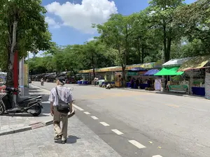 Bối cảnh là một con đường tại thị xã Sơn Tây vào một ngày nắng đẹp, hai bên đường có nhiều cây xanh. Dọc bên phải đường là một dãy các gian hàng tạm được dựng lên, tạo thành một khu chợ hoặc hội chợ trưng bày sản phẩm. Hoạt động chính là mua bán và tham quan; có nhiều người đang tụ tập tại các gian hàng, một số người đang đi bộ. Ở phía trước, một người đàn ông lớn tuổi đang đi bộ trên vỉa hè. Các vật thể có trong ảnh bao gồm xe máy, các gian hàng có mái che, bàn ghế, và nhiều loại hàng hóa, nổi bật nhất là những đống sầu riêng lớn. Có hai người phụ nữ đội nón lá đang đi bộ gần các gian hàng.。Các dòng chữ ghi nhận được từ các biển hiệu và băng rôn:。- Băng rôn lớn màu vàng: "KHU TRƯNG BÀY, GIỚI THIỆU SẢN PHẨM ĐẶC TRƯNG THỊ XÃ SƠN TÂY" và logo "OCOP".。- Biển quảng cáo sự kiện: "SỰ KIỆN QUẢNG BÁ VÀ TÔN VINH CÁC SẢN PHẨM OCOP... PHẨM VỀ MÍT... HÀ NỘI NĂM 2024".。- Biển hiệu gian hàng: "Khoamộc DECOR - COFFEE".。- Biển hiệu gian hàng khác: "HỢP TÁC XÃ TƯƠNG LÀNG BỘ PHƯƠNG VIỆT ĐẶC SẢN TRUYỀN THỐNG" và "TƯƠNG LÀNG BỘ".。（日本語訳）。背景は、晴れた日のソンタイ市社のある道で、道の両側には多くの緑の木があります。道の右側に沿って、製品を展示する市場やフェアを形成している一列の仮設の屋台が建てられています。主な活動は売買と見物です。多くの人々が屋台に集まっており、歩いている人もいます。前方には、一人の年配の男性が歩道を歩いています。写真にある物体には、バイク、屋根付きの屋台、テーブルと椅子、そして多くの種類の商品が含まれ、最も目立つのは大きなドリアンの山です。ノンラーをかぶった二人の女性が屋台の近くを歩いています。看板や横断幕から読み取れる文字：。- 大きな黄色の横断幕：「KHU TRƯNG BÀY, GIỚI THIỆU SẢN PHẨM ĐẶC TRƯNG THỊ XÃ SƠN TÂY」（ソンタイ市社特産品展示・紹介エリア）と「OCOP」のロゴ。- イベント広告看板：「SỰ KIỆN QUẢNG BÁ VÀ TÔN VINH CÁC SẢN PHẨM OCOP... PHẨM VỀ MÍT... HÀ NỘI NĂM 2024」（OCOP製品の広報と顕彰イベント...ジャックフルーツ製品...ハノイ2024年）。- 屋台の看板：「Khoamộc DECOR - COFFEE」。- 別の屋台の看板：「HỢP TÁC XÃ TƯƠNG LÀNG BỘ PHƯƠNG VIỆT ĐẶC SẢN TRUYỀN THỐNG」（ボー村の味噌協同組合 伝統特産品）と「TƯƠNG LÀNG BỘ」（ボー村の味噌）。