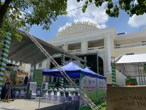 Bức ảnh chụp tại khu vực ngoài trời phía trước Trung tâm Văn hóa Thể thao Sơn Tây. Trên nóc tòa nhà có dòng chữ vàng "TRUNG TÂM VĂN HÓA THỂ THAO SƠN TÂY". Khung cảnh cho thấy sự chuẩn bị cho một sự kiện, với một sân khấu lớn có khung giàn kim loại và mái che đang được dựng lên. Phía trước sân khấu, nhiều hàng ghế được phủ vải trắng và thắt nơ màu xanh lá cây được sắp xếp cho khán giả. Xung quanh có các nhà bạt màu xanh, quạt điều hòa và các cột trang trí sọc xanh trắng. Các biểu ngữ quảng cáo cho sự kiện được treo lên, có thể đọc được nội dung: "SỰ KIỆN QUẢNG BÁ SẢN PHẨM OCOP CÁC TỈNH ĐỒNG BẰNG SÔNG HỒNG" và "GIAN HÀNG TRƯNG BÀY, GIỚI THIỆU VÀ BÁN SẢN PHẨM OCOP THÀNH PHỐ HÀ NỘI NĂM 2024", cùng với logo OCOP. Ngoài ra còn có một vật trang trí bằng tre treo những chiếc nơm (dụng cụ bắt cá truyền thống). Ở tiền cảnh có một tấm bia đá khắc chữ đỏ "TRUNG TÂM V... THỂ TH...". Thời tiết nắng đẹp, trời trong xanh có mây.。（日本語訳）。写真はソンタイ文化スポーツセンター前の屋外エリアで撮影されたものです。建物の屋上には「TRUNG TÂM VĂN HÓA THỂ THAO SƠN TÂY」という黄色い文字があります。この光景は、金属のトラスフレームと屋根を持つ大きなステージが設営されており、あるイベントの準備が進められていることを示しています。ステージの前には、白い布で覆われ緑色のリボンが結ばれた椅子が何列も観客のために並べられています。周りには青いテント、冷風扇、そして青と白の縞模様の装飾柱があります。イベントを宣伝する横断幕が掲げられており、「SỰ KIỆN QUẢNG BÁ SẢN PHẨM OCOP CÁC TỈNH ĐỒNG BẰNG SÔNG HỒNG」（紅河デルタ各省OCOP製品プロモーションイベント）および「GIAN HÀNG TRƯNG BÀY, GIỚI THIỆU VÀ BÁN SẢN PHẨM OCOP THÀNH PHỐ HÀ NỘI NĂM 2024」（2024年ハノイ市OCOP製品展示・紹介・販売ブース）という内容が、OCOPのロゴと共に読み取れます。その他に、ノム（伝統的な魚を捕る道具）を吊るした竹製の装飾物もあります。前景には「TRUNG TÂM V... THỂ TH...」と赤い文字が刻まれた石碑があります。天気は晴れで、空は青く澄み、雲があります。