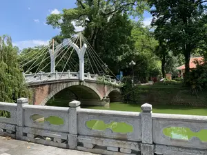 Bức ảnh chụp một cây cầu cổ có kiến trúc độc đáo bắc qua một con hào nước, có khả năng là trong một khu di tích lịch sử hoặc công viên. Cây cầu được xây theo kiểu vòm bằng gạch, phía trên có kết cấu dây văng trang trí màu trắng. Lan can cầu và lan can ở tiền cảnh đều được làm bằng đá và có chạm khắc hoa văn. Nước trong hào có màu xanh lục. Xung quanh cầu là không gian với nhiều cây cổ thụ to lớn, xanh tốt. Bầu trời trong xanh, có nắng và vài gợn mây trắng. Phía xa bên kia cầu có thể nhìn thấy một phần của một công trình kiến trúc mái ngói và một bảng thông tin nhưng không thể đọc rõ chữ. Khung cảnh yên bình, không có hoạt động của con người.。（日本語訳）。写真には、歴史的遺跡または公園の中にあると思われる、水堀にかかるユニークな建築様式の古い橋が写っている。橋はレンガ造りのアーチ様式で建てられており、上部には白い装飾的な斜張構造がある。橋の欄干と手前の欄干は、いずれも石でできており、模様が彫られている。堀の水は緑色である。橋の周りは、大きく青々とした多くの古木がある空間である。空は青く澄み、日が差しており、白い雲がいくつか浮かんでいる。平和な光景で、人の活動は見られない。