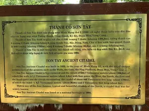 Bức ảnh chụp một tấm bia thông tin bằng kim loại màu vàng đồng tại khu di tích Thành cổ Sơn Tây. Tấm bia được đặt trên một bệ đá, có khắc chữ màu đen bằng cả tiếng Việt và tiếng Anh để giới thiệu về lịch sử và kiến trúc của thành cổ. Xung quanh tấm bia có khung viền hoa văn trang trí.。Nội dung trên bia:。THÀNH CỔ SƠN TÂY。Thành cổ Sơn Tây được xây dựng năm Minh Mạng thứ 3 (1822) với nghệ thuật kiến trúc độc đáo của các hạng mục như: Tường thành, Cổng thành, Kỳ đài, Đoan Môn, Vọng Cung...。Thành cổ Sơn Tây hình tứ giác với chu vi 326 trượng 7 thước (khoảng 1306,8m), tường thành cao 1 trượng 1 thước (khoảng 4,4m), 4 cổng thành: Tây, Bắc, Đông, Nam. Chu vi hào nước bao quanh thành là 448 trượng (khoảng 1792m), rộng 6 trượng 7 thước (khoảng 26,8m), sâu 1 trượng (khoảng 4m).。Thành cổ Sơn Tây là một trong bốn tòa thành nổi tiếng, tiêu biểu và đẹp nhất Bắc Kỳ, đã đi vào lịch sử và được xếp hạng di tích lịch sử quốc gia năm 1994.。SON TAY ANCIENT CITADEL。Son Tay Ancient Citadel was built in 1822, in the year of Minh Mang III, with the art of unique architecture of the projects such as the Walls, the Gate, the Flag Platform, Doan Mon, Vong Cung ...。Son Tay Ancient Citadel is four-cornered and the circuit of 326,7 Vietnamese metres (about 1306,8m), of which the walls is 1,1 Vietnamese metres (about 4,4m) with four gates: the West, the North, the East and the South gates and the circuit surrounding the citadel of about 448 Vietnamese feet long (about 1792m), of which the width is 6,7 Vietnamese metres (about 26,8m) and 1 Vietnamese foot deep (about 4m).。This is one of the four famous, typical and beautiful citadels of the North, a citadel that was far widely known.。Son Tay Ancient Citadel was listed as a national heritage in 1994.。（日本語訳）。ソンタイ（山西）古城遺跡にある、銅色の金属製の案内板を写した写真。案内板は石の台座の上に置かれ、古城の歴史と建築を紹介するために、ベトナム語と英語の両方で黒い文字が刻まれている。案内板の周りには、装飾的な模様の縁取りがある。碑文の内容：。ソンタイ（山西）古城。ソンタイ古城は、ミンマン（明命）帝3年（1822年）に、城壁、城門、旗台、端門、望宮などの各項目における独特な建築芸術をもって建設された。ソンタイ古城は四角形で、周囲は326丈7尺（約1306.8m）、城壁の高さは1丈1尺（約4.4m）で、西、北、東、南の4つの城門がある。城を囲む堀の周囲は448丈（約1792m）、幅は6丈7尺（約26.8m）、深さは1丈（約4m）である。ソンタイ古城は、北圻（トンキン）で最も有名で、代表的かつ美しい4つの城の一つであり、歴史に名を刻み、1994年に国家歴史遺跡に指定された。ソンタイ古城。ソンタイ古城は、ミンマン帝3年（1822年）に、城壁、城門、旗台、端門、望宮などの独特な建築様式で建設された。ソンタイ古城は四角形で、周囲は326.7ベトナムメートル（約1306.8m）、城壁は1.1ベトナムメートル（約4.4m）で、西、北、東、南の4つの門がある。城を囲む周囲は約448ベトナムフィート（約1792m）で、その幅は6.7ベトナムメートル（約26.8m）、深さは1ベトナムフィート（約4m）である。これは北部で有名、代表的かつ美しい4つの城の一つであり、広く知られた城であった。ソンタイ古城は1994年に国の遺産として登録された。