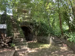 Bức ảnh ghi lại một khung cảnh tại một địa điểm có vẻ là di tích lịch sử, có thể là một phần của một thành cổ. Nổi bật là một công trình kiến trúc bằng đá hoặc bê tông đã cũ kỹ, với một lối vào hình vòm tối tăm. Hai bên là hai cầu thang ngoài trời, cũng làm bằng vật liệu tương tự và đã phủ đầy rêu xanh cùng cây cỏ dại, cho thấy dấu vết của thời gian. Cầu thang bên trái dài hơn, dẫn lên phần trên của công trình, trong khi cầu thang bên phải ngắn hơn. Cảnh vật xung quanh rất um tùm với nhiều cây lớn, lùm tre và cây dây leo bao phủ các bức tường. Sân được lát gạch cũ, lốm đốm rêu và bóng cây. Dưới chân cầu thang bên trái có một đống củi gỗ. Không có người hay hoạt động nào diễn ra trong ảnh. Không có văn bản hay biển báo nào có thể nhìn thấy rõ.。（日本語訳）。写真は、歴史的遺跡と思われる場所、おそらくは古い城の一部の光景を捉えています。目立っているのは、石かコンクリートでできた古びた建築物で、暗いアーチ型の入口があります。両側には2つの屋外階段があり、それらも同様の素材で作られ、緑の苔や雑草で覆われており、時の痕跡を示しています。左側の階段はより長く、建築物の上部へと続いていますが、一方、右側の階段はより短いです。周囲の景色は、多くの大きな木々、竹やぶ、そして壁を覆うつる植物で非常に生い茂っています。中庭は古いレンガで舗装されており、苔が点在し、木々の影が落ちています。左側の階段の麓には、薪の山があります。写真の中には人や何の活動も見られません。