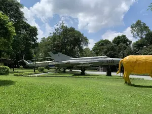 Bối cảnh là một khu trưng bày ngoài trời, có thể là một công viên hoặc bảo tàng, vào một ngày nắng đẹp. Đối tượng chính là một chiếc máy bay chiến đấu phản lực màu bạc, mang số hiệu "5238" màu đỏ, được đặt trên bệ đỡ trên một khu đất có cỏ. Dưới cánh máy bay có gắn các tên lửa. Ở phía sau, có thể nhìn thấy một phần của một chiếc trực thăng mang số hiệu "7822" và dòng chữ "KHÔNG QUÂN VIỆT NAM". Trên đuôi của chiếc máy bay chiến đấu cũng có dòng chữ "KHÔNG QUÂN VIỆT NAM" nhưng hơi mờ. Bên phải tiền cảnh là phần sau của một bức tượng lớn màu vàng, có hình dáng một con vật giống như con trâu hoặc con bò. Xung quanh là không gian xanh với bãi cỏ, cây cối rậm rạp và lối đi bộ. Bầu trời trong xanh với những đám mây trắng. Không có người hay hoạt động nào được ghi nhận, chỉ là cảnh vật tĩnh tại một khu trưng bày.。（日本語訳）。背景は、晴れた日の屋外展示エリアで、公園または博物館のようです。主な対象は、草の生えた土地の台座の上に置かれた、赤い番号「5238」を付けた銀色のジェット戦闘機です。飛行機の翼の下にはミサイルが取り付けられています。後方には、番号「7822」と「KHÔNG QUÂN VIỆT NAM」の文字が入ったヘリコプターの一部が見えます。戦闘機の尾翼にも「KHÔNG QUÂN VIỆT NAM」の文字がありますが、少しぼやけています。前景の右側には、水牛か牛のような動物の形をした、大きな黄色い像の後部があります。周囲は芝生、生い茂った木々、遊歩道のある緑の空間です。空は青く、白い雲が浮かんでいます。人や活動は見られず、展示エリアの静かな光景があるだけです。