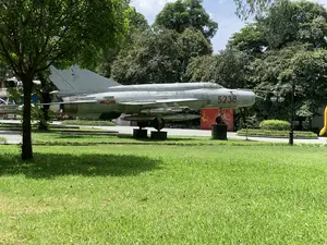 Bức ảnh chụp một chiếc máy bay chiến đấu được trưng bày ngoài trời trên một bãi cỏ xanh. Đây là một chiếc máy bay phản lực màu xám, có số hiệu "5238" được sơn màu đỏ ở phần mũi. Trên thân máy bay có biểu tượng ngôi sao vàng năm cánh trên nền tròn màu đỏ, là phù hiệu của Không quân Nhân dân Việt Nam. Máy bay được đặt trên các bệ đỡ và có trang bị tên lửa dưới cánh. Khung cảnh xung quanh là một công viên hoặc khu bảo tàng với bãi cỏ rộng, nhiều cây xanh và một cột đèn đường. Phía sau máy bay có một tấm phông lớn màu đỏ với họa tiết trang trí màu vàng.。（日本語訳）。これは灰色のジェット機で、機首部分に「5238」という機体番号が赤色で塗装されています。機体には、赤い円形の背景に五芒星の金色の星が描かれた、ベトナム人民空軍の記章があります。航空機は台座の上に置かれており、翼の下にはミサイルが装備されています。周囲の風景は、広大な芝生、多くの緑の木々、そして一本の街灯がある公園または博物館エリアです。航空機の後ろには、黄色の装飾模様が入った大きな赤色の背景幕があります。