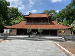 Bức ảnh chụp một ngôi đền hoặc đình cổ kính của Việt Nam, có khả năng là một di tích lịch sử tại khu vực Sơn Tây. Công trình chính có kiến trúc truyền thống với hai tầng mái ngói đỏ, các góc mái cong vút được trang trí hình linh vật. Tòa nhà được xây dựng chủ yếu bằng gỗ và gạch, nằm trên một nền đá cao. Phía trước là một khoảng sân rộng được lát đá phiến màu xám. Chính giữa sân, phía trước các bậc thang, là một lư hương lớn bằng đá. Lối lên đền là các bậc tam cấp bằng đá, với lan can được chạm khắc hình nghê đá. Ở hai bên sân có cắm cờ hội nhiều màu sắc. Trên tường gạch phía bên phải có một tấm bia đá nhưng không thể đọc rõ nội dung. Xung quanh ngôi đền là rất nhiều cây xanh cổ thụ, tạo nên một không gian trang nghiêm và yên tĩnh dưới bầu trời xanh trong. Không có hoạt động nào của con người được ghi nhận trong ảnh.。（日本語訳）。写真はベトナムの古風な寺院または亭を写しており、ソンタイ地区にある歴史的遺跡である可能性があります。主な建物は伝統的な建築様式で、赤い瓦の二層の屋根を持ち、湾曲して反り上がった屋根の角は霊獣の像で装飾されています。建物は主に木とレンガで建てられており、高い石の土台の上にあります。前には灰色のスレートで舗装された広い庭があります。庭の中央、階段の前には、石でできた大きな香炉があります。寺院への通路は石の三段の階段で、欄干には石の獅子（Nghê）が彫刻されています。庭の両側には、色とりどりの祭りの旗が立てられています。右側のレンガの壁には石碑がありますが、内容ははっきりと読み取れません。寺院の周りには多くの古い緑の木々があり、澄んだ青空の下で荘厳で静かな空間を作り出しています。写真には人間の活動は記録されていません。