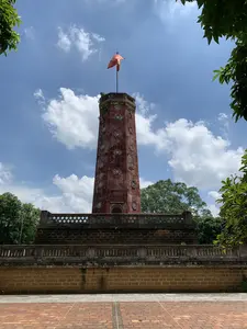 Hình ảnh chụp một tòa tháp cổ, đây là Cột cờ Sơn Tây, một di tích lịch sử. Công trình có kiến trúc hình bát giác, được xây trên một bệ cao bằng gạch đá ong. Thân tháp có màu đỏ sẫm, mang dấu ấn thời gian, và được trang trí bằng các cửa sổ hình tròn cách điệu. Trên đỉnh tháp có một lá cờ màu cam đang tung bay dưới bầu trời xanh có nhiều mây trắng. Xung quanh chân tháp là lan can với các hoa văn trang trí. Phía trước là một khoảng sân rộng lát gạch. Cây cối xanh tươi bao quanh khu vực. Không có hoạt động của con người được ghi lại, khung cảnh tĩnh lặng.。（日本語訳）。古い塔を撮影した写真、これは歴史的遺跡であるソンタイの旗竿です。この建造物は八角形の建築様式で、ラテライト煉瓦の高い台座の上に建てられています。塔の本体は濃い赤色で、時の痕跡を帯びており、様式化された円形の窓で装飾されています。塔の頂上には、白い雲が多く浮かぶ青空の下で、オレンジ色の旗がはためいています。塔の麓の周りには、装飾的な模様が施された手すりがあります。前方には、レンガ敷きの広い中庭があります。緑豊かな木々がそのエリアを囲んでいます。人の活動は記録されておらず、静かな光景です。