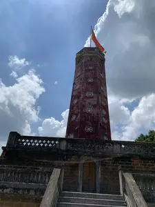 Bức ảnh chụp một công trình kiến trúc cổ, nhìn từ góc thấp hướng lên trên. Đây là Kỳ đài nằm trong khu di tích Thành cổ Sơn Tây. Tòa tháp chính có dạng hình bát giác, được xây bằng gạch cũ màu đỏ sẫm, trên thân có nhiều họa tiết trang trí hình bánh xe. Trên đỉnh tháp, lá cờ đỏ sao vàng của Việt Nam đang tung bay trên nền trời xanh và mây trắng. Tháp được đặt trên một bệ đá vững chãi, xây bằng đá ong đặc trưng. Xung quanh bệ có lan can với các ô thoáng hình chữ thập. Một cầu thang bằng đá dẫn lên phía trên, nơi có một lối vào với cửa vòm. Không có chữ viết nào có thể đọc được trong hình.。（日本語訳）。古代の建築物を下からのアングルで見上げるように撮影した写真です。これはソンタイ（Sơn Tây）古城遺跡内にあるフラッグタワー（Kỳ đài）です。主塔は八角形で、暗赤色の古いレンガで建てられており、塔身には車輪の形をした装飾模様が多くあります。塔の頂上では、ベトナムの金星紅旗が青い空と白い雲を背景にはためいています。塔は、特徴的なラテライトで造られた頑丈な石の台座の上に建てられています。台座の周りには、十字形の通気口がある欄干があります。石の階段が上へと続いており、そこにはアーチ型の扉がある入り口があります。写真の中には読み取れる文字はありません。