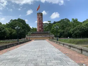 Bức ảnh chụp một di tích lịch sử vào một ngày trời trong xanh, có mây trắng. Trung tâm của bức ảnh là Cột cờ Thành cổ Sơn Tây, một công trình kiến trúc cổ kính. Cột cờ có dạng hình trụ bát giác, được xây bằng gạch màu đỏ, trên đỉnh có lá cờ đỏ sao vàng của Việt Nam đang tung bay. Thân cột cờ có các ô cửa sổ trang trí hình tròn. Cột cờ được đặt trên một bệ đá cao, nhiều tầng. Có một tấm bia khắc chữ trên thân cột cờ nhưng không thể đọc rõ nội dung. Một con đường rộng được lát bằng đá phiến màu xám ở giữa và gạch đỏ ở hai bên dẫn thẳng đến chân cột cờ. Dọc hai bên lối đi là các cột đèn đường kiểu cổ. Bao quanh khu di tích là rất nhiều cây xanh tươi tốt. Khung cảnh vắng lặng, trang nghiêm, không có hoạt động nào của con người được ghi nhận.。（日本語訳）。写真は、白い雲が浮かぶ青空の日に撮影された史跡です。写真の中心には、古い建築物であるソンタイ古城の旗竿があります。旗竿は八角形の円柱状で、赤レンガで造られており、頂上にはベトナムの金星紅旗がはためいています。旗竿の本体には、円形の装飾的な窓があります。旗竿は、高く多層の石の台座の上に置かれています。旗竿の本体には文字が刻まれた石碑がありますが、内容ははっきりと読み取れません。中央が灰色の石板で、両側が赤レンガで舗装された広い道が、旗竿の麓までまっすぐ続いています。通路の両側には、古い様式の街灯が並んでいます。史跡の周りは、青々とした多くの木々に囲まれています。景色は静かで荘厳であり、人間の活動は見られません。