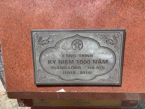 Hình ảnh chụp cận cảnh một tấm biển kỷ niệm bằng kim loại gắn trên bệ đá hoa cương màu đỏ. Tấm biển này là một công trình kỷ niệm 1000 năm Thăng Long - Hà Nội. Nội dung được khắc trên biển như sau:。Phần trên cùng là biểu tượng Khuê Văn Các với chữ "HÀ NỘI" bên dưới.。Nội dung chính:。"CÔNG TRÌNH。KỶ NIỆM 1000 NĂM。THĂNG LONG - HÀ NỘI。(1010 - 2010)"。Tấm biển được trang trí bằng các họa tiết hoa văn truyền thống như mây và sóng nước ở các góc. Bề mặt kim loại của tấm biển có nhiều vết xước và hình vẽ bậy.。Bên dưới tấm biển chính, có một tấm biển nhỏ hơn màu vàng ghi tên đơn vị tặng, có thể đọc được chữ "HANDICO" và "KÍNH TẶNG".。（日本語訳）。赤い花崗岩の台座に取り付けられた金属製の記念プレートのクローズアップ写真。このプレートは、タンロン・ハノイ遷都1000年を記念する事業のものです。プレートに刻まれた内容は以下の通りです。最上部は奎文閣（クエ・ヴァン・カック）のシンボルで、その下に「HÀ NỘI」の文字があります。主な内容：。「タンロン・ハノイ遷都1000年記念事業。(1010 - 2010)」。プレートは、角に雲や波などの伝統的な文様で装飾されています。プレートの金属表面には、多くの傷や落書きがあります。メインプレートの下には、寄贈した団体の名前が記された小さな黄色のプレートがあり、「HANDICO」と「謹贈」の文字が読み取れます。