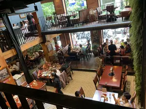 Bối cảnh là bên trong một nhà hàng có không gian rộng rãi, được thiết kế theo kiểu có gác lửng hoặc nhiều tầng, được chụp từ trên cao nhìn xuống. Rất đông khách hàng, bao gồm cả người lớn và trẻ em, đang ngồi ăn uống và trò chuyện tại các bàn. Nhân viên phục vụ đang di chuyển để phục vụ khách. Nội thất của nhà hàng có bàn ghế gỗ, tường gạch, và sàn lát gạch hoa văn. Một số bàn được trang bị bếp lẩu. Nhìn qua các ô cửa kính lớn, có thể thấy quang cảnh đường phố bên ngoài với nhiều xe máy đang đậu. Có một biển báo "Cấm hút thuốc" (NO SMOKING) trên cột gỗ và một biển báo cấm hút thuốc khác trên cột gạch.。（日本語訳）。背景は、中二階または多層階建てに設計された広々とした空間のレストランの店内で、上から見下ろすように撮影されている。大人と子供を含む非常に多くの客が、テーブルで食事をしたり会話をしたりしている。接客スタッフが客にサービスを提供するために動き回っている。レストランの内装には、木製のテーブルと椅子、レンガの壁、模様のあるタイル張りの床がある。いくつかのテーブルには鍋用のコンロが備え付けられている。木製の柱に「禁煙」（NO SMOKING）の標識が一つあり、レンガの柱にもう一つの禁煙の標識がある。