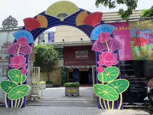 Bối cảnh là lối vào của Khu di sản Hoàng thành Thăng Long ở Hà Nội. Một cổng chào lớn hình vòm được dựng lên, trang trí rực rỡ với các họa tiết quạt giấy và hoa sen nhiều màu sắc, dường như để chào mừng một sự kiện. Phía sau cổng chào là lối vào tòa nhà, nơi có quầy bán vé.。Các thông tin bằng chữ viết có thể đọc được bao gồm:。- Trên biển hiệu chính của tòa nhà: 'NƠI BÁN VÉ', 'ĐÓN TIẾP KHÁCH THAM QUAN', 'TICKET OFFICE', 'RECEPTION AND INFORMATION SECTION', 'DI SẢN VĂN HÓA THẾ GIỚI KHU TRUNG TÂM HOÀNG THÀNH THĂNG LONG HÀ NỘI', 'CENTRAL SECTOR OF THE IMPERIAL CITADEL OF THANG LONG'.。- Trên các biểu ngữ và áp phích quảng cáo sự kiện: 'Tết Đoan Ngọ', 'MẠC TRƯNG BÀY:', 'EXPOSITION | EXHIBITION', '...au musée parcours d'objets', 'hành trình của hiện vật', 'museum: route of objects', cùng với thông tin về các đơn vị đồng tổ chức như 'Tòa Thị chính Thành phố Toulouse' và 'Cơ quan phát triển Pháp - AFD'.。- Trên các biểu ngữ dọc: 'THĂNG LONG IMPERIAL CITADEL'.。- Trên chậu cây bằng xi măng: 'WELCOME'.。Địa điểm này là lối vào một khu di tích lịch sử, cụ thể là Hoàng thành Thăng Long. Hoạt động đang diễn ra là việc chuẩn bị và trang trí để đón khách tham quan cho một sự kiện văn hóa là 'Tết Đoan Ngọ' và một cuộc triển lãm. Các vật thể chính trong ảnh bao gồm cổng chào trang trí, các biển hiệu, tòa nhà bán vé, chậu cây, và một phần chiếc xe máy đang đỗ ở bên phải.。（日本語訳）。背景はハノイにあるタンロン皇城遺跡地区の入り口です。イベントを祝うためか、大きなアーチ型の歓迎ゲートが建てられ、色とりどりの紙扇子や蓮の花のモチーフで華やかに装飾されています。歓迎ゲートの後ろには、チケット売り場のある建物の入り口があります。読み取れる文字情報は以下の通りです：。- 建物のメイン看板：「NƠI BÁN VÉ」（チケット販売所）、「ĐÓN TIẾP KHÁCH THAM QUAN」（来場者受付）、「TICKET OFFICE」、「RECEPTION AND INFORMATION SECTION」、「DI SẢN VĂN HÓA THẾ GIỚI KHU TRUNG TÂM HOÀNG THÀNH THĂNG LONG HÀ NỘI」（世界文化遺産ハノイ・タンロン皇城中心地区）、「CENTRAL SECTOR OF THE IMPERIAL CITADEL OF THANG LONG」。- イベントを宣伝する横断幕やポスター：「Tết Đoan Ngọ」（端午の節句）、「MẠC TRƯNG BÀY:」（展示）、「EXPOSITION | EXHIBITION」、「...au musée parcours d'objets」（博物館にて、オブジェの旅）、「hành trình của hiện vật」（現物の旅）、「museum: route of objects」、そして「Tòa Thị chính Thành phố Toulouse」（トゥールーズ市役所）や「Cơ quan phát triển Pháp - AFD」（フランス開発庁 - AFD）といった共同主催者に関する情報。- 縦型の横断幕：「THĂNG LONG IMPERIAL CITADEL」。- セメント製の植木鉢：「WELCOME」。この場所は歴史遺跡地区、具体的にはタンロン皇城への入り口です。現在行われている活動は、「Tết Đoan Ngọ」（端午の節句）という文化イベントと展示会のために来場者を迎える準備と装飾です。写真に写っている主な物体には、装飾された歓迎ゲート、各種看板、チケット販売所のある建物、植木鉢、そして右側に駐車しているバイクの一部が含まれます。