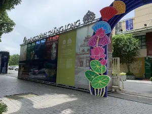 Bối cảnh là một khu vực trưng bày ngoài trời, có thể là lối vào của Khu di tích Hoàng thành Thăng Long. Một cổng chào lớn được dựng lên trên vỉa hè lát gạch. Trên đỉnh cổng có dòng chữ lớn bằng kim loại "HOÀNG THÀNH THĂNG LONG". Thân cổng được tạo thành từ nhiều tấm pa-nô quảng cáo lớn, trên đó có các thông tin và hình ảnh về di tích.。Nội dung văn bản có thể đọc được bao gồm:。- Trên pa-nô màu xanh lá: "DI SẢN VĂN HÓA THẾ GIỚI KHU TRUNG TÂM HOÀNG THÀNH THĂNG LONG - HÀ NỘI", "THE CENTRAL SECTOR OF THE IMPERIAL CITADEL OF THANG LONG, HANOI", và logo "HOÀNG THÀNH THĂNG LONG" cùng logo Di sản Thế giới của UNESCO với ghi chú "World Heritage since 2010".。- Trên các pa-nô khác: "GLAZED TERRACOTA", "CELADON GLAZED LION", "WORLD CULTURAL HERITAGE SITE", "TERRACOTTA PHOENIX HEAD", "YELLOW TUBULAR TILES".。- Một biển báo riêng ở bên trái ghi: "19C HOÀNG ĐIỆU", "PERMANENT EXHIBITION".。- Các biểu ngữ treo trên tòa nhà phía sau: "THĂNG LONG IMPERIAL CITADEL".。Bên cạnh các pa-nô là các vật trang trí lớn, đầy màu sắc hình hoa sen, lá sen và những chiếc quạt giấy. Khung cảnh xung quanh có cây xanh, một phần cổng sắt và một tòa nhà. Không có hoạt động nào của con người được ghi lại, đây là một cảnh tĩnh giới thiệu về khu di tích.。（日本語訳）。背景は屋外の展示エリアで、タンロン皇城遺跡地区の入り口である可能性があります。レンガ敷きの歩道の上に大きな歓迎ゲートが建てられています。ゲートの上部には「HOÀNG THÀNH THĂNG LONG」という大きな金属製の文字があります。ゲート本体は多くの大きな広告パネルで構成されており、そこには遺跡に関する情報や画像が掲載されています。読み取れるテキストの内容は以下の通りです：。- 緑色のパネルには：「DI SẢN VĂN HÓA THẾ GIỚI KHU TRUNG TÂM HOÀNG THÀNH THĂNG LONG - HÀ NỘI」、「THE CENTRAL SECTOR OF THE IMPERIAL CITADEL OF THANG LONG, HANOI」、そして「HOÀNG THÀNH THĂNG LONG」のロゴと、「World Heritage since 2010」という注記付きのユネスコ世界遺産のロゴ。- 他のパネルには：「GLAZED TERRACOTA」、「CELADON GLAZED LION」、「WORLD CULTURAL HERITAGE SITE」、「TERRACOTTA PHOENIX HEAD」、「YELLOW TUBULAR TILES」。- 左側にある別の看板には、「19C HOÀNG ĐIỆU」、「PERMANENT EXHIBITION」と書かれています。- 後ろの建物に掲げられた横断幕には：「THĂNG LONG IMPERIAL CITADEL」。パネルの隣には、蓮の花、蓮の葉、そして紙の扇の形をした、大きくてカラフルな装飾品があります。周囲の風景には、緑の木々、鉄の門の一部、そして建物があります。人間の活動は記録されておらず、これは遺跡地区を紹介する静的な光景です。