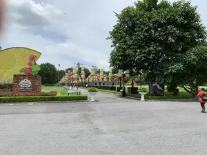 Bối cảnh là một con đường dẫn vào khu di tích Hoàng thành Thăng Long. Ở bên trái có một tấm biển xây bằng gạch với logo và dòng chữ "IMPERIAL CITADEL OF THANG LONG". Phía sau tấm biển là một vật trang trí lớn hình chiếc quạt màu vàng có họa tiết hoa sen. Con đường được trải nhựa, hai bên là bãi cỏ xanh và cây cối. Dọc theo con đường, có nhiều dãy đèn lồng màu đỏ và vàng được treo trên các cột. Một vài người đang đi dạo trên con đường, có thể là khách tham quan. Ở phía xa, có thể nhìn thấy một phần của bức tường thành cổ. Bầu trời có mây và có một chiếc máy bay đang bay.。（日本語訳）。背景は、タンロン城遺跡地区へと続く道です。左側には、ロゴと「IMPERIAL CITADEL OF THANG LONG」という文字が書かれた、レンガ造りの看板があります。看板の後ろには、蓮の花の模様がある黄色い扇形の大きな装飾物があります。道は舗装されており、両側には緑の芝生と木々があります。道に沿って、柱に吊るされた赤と黄色の提灯がいくつも並んでいます。何人かの人々が道を散歩しており、おそらく観光客でしょう。遠くには、古い城壁の一部が見えます。空は曇っており、飛行機が一機飛んでいます。