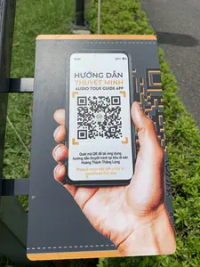 Bức ảnh chụp một tấm biển chỉ dẫn đặt ngoài trời, trên nền cỏ xanh. Tấm biển này cung cấp thông tin về ứng dụng hướng dẫn tham quan tự động bằng âm thanh cho du khách. Trên biển có in hình một bàn tay đang cầm điện thoại thông minh, màn hình hiển thị một mã QR để tải ứng dụng. Nội dung văn bản trên biển bao gồm:。- Tiêu đề tiếng Việt: "HƯỚNG DẪN THUYẾT MINH"。- Tiêu đề tiếng Anh: "AUDIO TOUR GUIDE APP"。- Hướng dẫn bằng tiếng Việt: "Quét mã QR để tải ứng dụng hướng dẫn thuyết minh tại khu di sản Hoàng Thành Thăng Long"。- Hướng dẫn bằng tiếng Anh: "Please scan the QR code to download the app"。Địa điểm được xác định là khu di sản Hoàng Thành Thăng Long dựa trên nội dung của tấm biển.。（日本語訳）。この案内板は、観光客向けの自動音声ガイドツアーアプリに関する情報を提供しています。案内板には、スマートフォンを持つ手が印刷されており、その画面にはアプリをダウンロードするためのQRコードが表示されています。案内板のテキスト内容は以下の通りです。- ベトナム語のタイトル：「HƯỚNG DẪN THUYẾT MINH」。- 英語のタイトル：「AUDIO TOUR GUIDE APP」。- ベトナム語の説明：「Quét mã QR để tải ứng dụng hướng dẫn thuyết minh tại khu di sản Hoàng Thành Thăng Long」（タンロン皇城遺跡地区の解説ガイドアプリをダウンロードするにはQRコードをスキャンしてください）。- 英語の説明：「Please scan the QR code to download the app」（アプリをダウンロードするにはQRコードをスキャンしてください）。案内板の内容に基づき、場所はタンロン皇城遺跡地区であると特定されます。