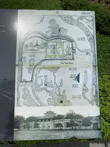 Bức ảnh chụp một bảng thông tin đặt ngoài trời trên nền cỏ xanh. Bảng thông tin này trưng bày một bản đồ cổ và một bức ảnh đen trắng cũ. Phần trên là bản đồ cổ của Hoàng thành Thăng Long, được vẽ bằng mực tàu với các ghi chú bằng chữ Hán. Bản đồ mô tả cấu trúc của thành cổ với các cung điện, hồ nước, và sông ngòi. Một số địa danh có thể nhận dạng bằng chữ Hán như: 北 (Bắc - Phía Bắc), 西湖 (Tây Hồ), 國子監 (Quốc Tử Giám), 寶天塔 (Báo Thiên Tháp), 奉天府 (Phụng Thiên Phủ), 司天監 (Tư Thiên Giám), và 東宮 (Đông Cung). Có một dòng chữ tiếng Anh "Kinh Thien Palace" (Điện Kính Thiên) chỉ vào một khu vực được đánh dấu màu xanh lá cây trên bản đồ. Phần dưới là một bức ảnh đen trắng chụp một công trình kiến trúc cổ, có thể là một cổng thành hoặc một phần của cung điện trong Hoàng thành, với tường thành và lầu gác. Bên trái của bảng thông tin có thể thấy một phần chữ tiếng Anh "...g Long -" và "... Centre". Đây là một bảng chỉ dẫn lịch sử, cung cấp thông tin về cấu trúc của Hoàng thành Thăng Long xưa cho du khách.。（日本語訳）。緑の芝生の上に屋外に設置された案内板を写した写真です。この案内板には、古い地図と古い白黒写真が展示されています。上部はタンロン皇城の古地図で、墨で描かれ、漢字で注釈が付けられています。地図は、宮殿、湖、川など、古い城の構造を描写しています。北（Bắc - 北）、西湖（Tây Hồ）、國子監（Quốc Tử Giám）、寶天塔（Báo Thiên Tháp）、奉天府（Phụng Thiên Phủ）、司天監（Tư Thiên Giám）、東宮（Đông Cung）など、いくつかの地名は漢字で識別できます。地図上で緑色にマークされた区域を指し示す、「Kinh Thien Palace」（敬天殿）という英語の文字列があります。下部は、古い建築物を写した白黒写真で、城壁と楼閣があり、おそらく城門か皇城内の宮殿の一部と思われます。案内板の左側には、「...g Long -」と「... Centre」という英語の文字の一部が見えます。これは歴史案内板であり、観光客に昔のタンロン皇城の構造に関する情報を提供しています。