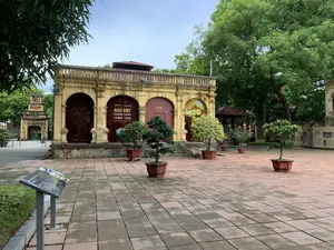 Bối cảnh là một khoảng sân rộng lát gạch bên trong khu di tích lịch sử Hoàng thành Thăng Long. Ở trung tâm là một tòa nhà kiến trúc cổ, tường sơn vàng, có các lối vào hình vòm. Một trong các lối vào có treo một biểu ngữ lớn với nội dung: "TRƯNG BÀY | EXHIBITION, BÁU VẬT HOÀNG CUNG THĂNG LONG, Treasures of Thang Long Imperial Palace". Phía trước tòa nhà, trên sân, có đặt một vài chậu cây cảnh bonsai. Bên trái phía trước có một bảng thông tin trên giá đỡ, có thể đọc được dòng chữ "Dragon Steps" và dòng chữ tiếng Việt phía trên có khả năng là "Điện Kính Thiên". Xung quanh khu vực là nhiều cây xanh lớn. Phía xa bên trái có thể thấy một công trình cổng cổ khác. Không có hoạt động nào của con người được ghi lại, khung cảnh yên tĩnh.。（日本語訳）。背景は、タンロン皇城歴史遺跡地区内にある、レンガ敷きの広い中庭です。中央には、壁が黄色に塗られ、アーチ型の入り口を持つ古い建築様式の建物があります。入り口の一つには、「展示 | EXHIBITION, タンロン皇宮の宝物, Treasures of Thang Long Imperial Palace」という内容の大きな横断幕が掲げられています。建物の前の中庭には、盆栽の鉢植えがいくつか置かれています。左手前にはスタンドに立てられた案内板があり、「Dragon Steps」という文字と、その上のベトナム語の文字がおそらく「敬天殿」であることが読み取れます。周辺には大きな緑の木がたくさんあります。左奥には、別の古い門の建造物が見えます。人の活動は見られず、静かな光景です。