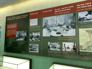 Đây là hình ảnh chụp một bảng trưng bày trong bảo tàng, ghi lại các sự kiện lịch sử quan trọng trong Chiến dịch Hồ Chí Minh vào tháng 4 năm 1975. Bảng trưng bày có màu xanh lá cây và đỏ, chứa nhiều hình ảnh, bản đồ, tài liệu và chú thích bằng cả tiếng Việt và tiếng Anh.。**Nội dung chữ viết được ghi lại:**。*   Tiêu đề chính: “THẦN TỐC - TÁO BẠO - BẤT NGỜ - CHẮC THẮNG”.。*   Các mốc thời gian và sự kiện: "07/04/1975", "14/04/1975", "26/04 - 30/04/1975 CHIẾN DỊCH HỒ CHÍ MINH", "28/04/1975 GIẢI PHÓNG BIÊN HÒA".。*   Chú thích cho các hình ảnh và tài liệu: "Quân ủy Trung ương họp ngày 31/03/1975 quyết định...", "Đại tướng Võ Nguyên Giáp - Bí thư Quân ủy Trung ương ra bức một điện số 1574...", "Tại Tổng Hành dinh (Nhà D67), Bộ Chính trị quyết định đặt tên Chiến dịch Sài Gòn - Gia Định là Chiến dịch Hồ Chí Minh", "Bộ Chỉ huy Chiến dịch Hồ Chí Minh tại Sở Chỉ huy tiền phương, tháng 04/1975...", "Quân Giải phóng tấn công làm chủ Biên Hòa, sân bay Tân Sơn Nhất", "Bản đồ miền Nam Việt Nam đánh dấu các khu vực quân đội Giải phóng làm chủ (màu đỏ)...".。**Cảnh và hoạt động được mô tả:**。*   **Nơi chốn:** Các bức ảnh mô tả các phòng họp tại Tổng Hành dinh (Nhà D67) và Sở Chỉ huy tiền phương, nơi các quyết định quan trọng được đưa ra.。*   **Hoạt động:** Các bức ảnh đen trắng ghi lại các hoạt động đa dạng: các nhà lãnh đạo quân sự và chính trị như Đại tướng Võ Nguyên Giáp và Đại tướng Văn Tiến Dũng đang họp, chỉ đạo chiến dịch trên bản đồ quân sự; các đơn vị bộ đội cùng xe tăng và thuyền đang hành quân tiến về Sài Gòn; cảnh tấn công vào sân bay.。*   **Vật thể:** Các vật thể chính bao gồm các bức ảnh lịch sử, bản sao tài liệu (bức điện mật), bản đồ quân sự, xe tăng, thuyền, và máy bay chiến đấu. Phía dưới bảng trưng bày có một tủ kính được chiếu sáng.。（日本語訳）。これは、1975年4月のホーチミン作戦における重要な歴史的出来事を記録した、博物館の展示パネルを撮影した写真です。展示パネルは緑と赤を基調とし、多くの写真、地図、資料、そしてベトナム語と英語の両方によるキャプションが含まれています。**記録されている文字の内容：**。*   メインタイトル：「神速・大胆・奇襲・必勝」。*   年月日と出来事：「1975年4月7日」、「1975年4月14日」、「1975年4月26日～4月30日 ホーチミン作戦」、「1975年4月28日 ビエンホア解放」。*   写真と資料のキャプション：「中央軍事委員会は1975年3月31日に会議を開き決定…」、「ヴォー・グエン・ザップ大将 - 中央軍事委員会書記が電報第1574号を発令…」、「総司令部（D67号棟）にて、政治局はサイゴン-ザーディン作戦をホーチミン作戦と命名することを決定」、「1975年4月、前線指揮所におけるホーチミン作戦司令部…」、「解放軍がビエンホア、タンソンニャット空港を攻撃し制圧」、「解放軍が制圧した地域（赤色）を記した南ベトナムの地図…」。**描写されている場面と活動：**。*   **場所：**写真は、重要な決定が下された総司令部（D67号棟）と前線指揮所の会議室を描写しています。*   **活動：**白黒写真は多様な活動を記録しています。ヴォー・グエン・ザップ大将やヴァン・ティエン・ズン大将のような軍事・政治指導者たちが会議を開き、軍事地図上で作戦を指揮している様子、戦車や船と共にサイゴンへ進軍する部隊、空港への攻撃シーンなどです。*   **物体：**主な物体には、歴史的な写真、資料のコピー（機密電報）、軍事地図、戦車、船、戦闘機が含まれます。展示パネルの下には、照明付きのガラスケースがあります。