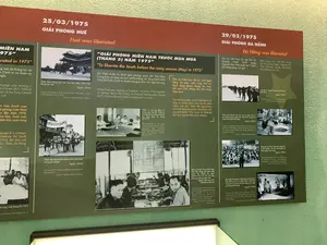 Đây là hình ảnh một bảng thông tin trưng bày trong bảo tàng, ghi lại các sự kiện lịch sử quan trọng trong Chiến dịch Mùa Xuân 1975. Bảng trưng bày được chia thành các phần, sử dụng các bức ảnh tư liệu đen trắng kèm theo chú thích bằng tiếng Việt và tiếng Anh để tường thuật về việc giải phóng Huế và Đà Nẵng.。Nội dung chi tiết trên bảng thông tin như sau:。1.  **Phần Giải phóng Huế (bên trái):**。*   Tiêu đề: "25/03/1975 GIẢI PHÓNG HUẾ".。*   Ảnh 1: Các binh sĩ Quân Giải phóng đang tiến vào cửa Ngọ Môn. Chú thích: "Quân Giải phóng tiến vào cửa Ngọ Môn, phía trước Thành nội Huế. Nguồn: TTXVN".。*   Ảnh 2: Người dân tuần hành trên đường phố để ăn mừng. Chú thích: "Nhân dân thành phố Huế mít tinh tuần hành mừng giải phóng. Nguồn: TTXVN". Một biểu ngữ trong ảnh ghi: "TOÀN DÂN ĐOÀN KẾT TÍCH CỰC".。2.  **Phần Giải phóng Đà Nẵng (bên phải):**。*   Tiêu đề: "29/03/1975 GIẢI PHÓNG ĐÀ NẴNG".。*   Văn bản: "Sau khi mất Huế, lại mất Tam Kỳ (Quảng Nam), chính quyền Nguyễn Văn Thiệu tuyên bố “tử thủ” Đà Nẵng bằng mọi giá."。*   Ảnh 1: Một hàng dài binh sĩ Quân Giải phóng đang diễu hành tiến vào thành phố. Chú thích: "Một đơn vị Quân Giải phóng tiến vào Đà Nẵng. Nguồn: TTXVN".。*   Ảnh 2: Quân Giải phóng cùng các phương tiện quân sự (xe tải, xe jeep) đã kiểm soát các cơ quan hành chính. Chú thích: "Quân Giải phóng làm chủ các cơ quan, đơn vị của chính quyền, quân đội Việt Nam Cộng hòa trong thành phố Đà Nẵng. Nguồn: TTXVN".。*   Ảnh 3: Hàng ngàn binh lính của quân đội Việt Nam Cộng hòa đang ngồi trên mặt đất sau khi ra trình diện chính quyền cách mạng. Chú thích: "Hàng vạn binh lính Việt Nam Cộng hòa ở tỉnh Quảng - Đà đã ra trình diện với chính quyền cách mạng. Nguồn: TTXVN".。*   Ảnh 4: Đại tướng Võ Nguyên Giáp đang nói chuyện với các sĩ quan trong một tòa nhà. Chú thích: "Đại tướng Võ Nguyên Giáp thăm cán bộ Quân Giải phóng tại trụ sở Quân đoàn 1 Việt Nam Cộng hòa tại thành phố Đà Nẵng (sau ngày giải phóng Đà Nẵng). Nguồn: TTXVN".。*   Ảnh 5: Một nhóm sĩ quan đang họp quanh một bàn có bản đồ. Trên bản đồ treo tường có chữ "HOA-YẾM".。3.  **Phần trung tâm (Bối cảnh và chỉ đạo chiến lược):**。*   Trích dẫn lớn: "“GIẢI PHÓNG MIỀN NAM TRƯỚC MÙA MƯA (THÁNG 5) NĂM 1975”".。*   Văn bản: "Khi Huế chuẩn bị được giải phóng, quân đội Việt Nam Cộng hòa rút chạy co cụm về Đà Nẵng, tại Tổng Hành dinh (Nhà D67) Bộ Chính trị nhanh chóng tổ chức Hội nghị mở rộng."。*   Văn bản: "Thời cơ chiến lược đã tới, một ngày bằng 20 năm, nắm vững thời cơ, hành động nhanh chóng, táo bạo, bất ngờ; Giải phóng Sài Gòn trước mùa mưa."。*   Ảnh 1: Các thành viên Bộ Chính trị đang họp trong một phòng họp, thể hiện sự vui mừng. Chú thích: "Niềm vui của các đồng chí trong cuộc họp Bộ Chính trị mở rộng khi tin thắng trận liên tục được báo về, tháng 03/1975".。*   Ảnh 2: Các lãnh đạo quân sự đang họp bàn. Chú thích: "Đồng chí Lê Đức Thọ và Đại tướng Văn Tiến Dũng phổ biến nghị quyết Bộ Chính trị ngày 25/03/1975 tại Sở Chỉ huy Quân giải phóng miền Nam. Nguồn: Bảo tàng LLVT miền Đông Nam Bộ".。（日本語訳）。これは博物館に展示されている案内板の画像で、1975年春季攻勢における重要な歴史的出来事を記録しています。展示パネルはいくつかのセクションに分かれており、ベトナム語と英語のキャプションが付いた白黒の資料写真を使用して、フエとダナンの解放について物語っています。案内板の詳細な内容は以下の通りです。1.  **フエ解放のセクション（左側）：**。*   見出し：「1975年3月25日 フエ解放」。*   写真1：解放軍の兵士たちが午門（ゴモン）に進軍している。キャプション：「解放軍がフエ城内の正面にある午門に進軍。出典：TTXVN（ベトナム通信社）」。*   写真2：市民が解放を祝って街頭を行進している。キャプション：「フエ市民が解放を祝って集会と行進を行う。出典：TTXVN」。写真の中の横断幕には「全人民団結積極」と書かれている。2.  **ダナン解放のセクション（右側）：**。*   見出し：「1975年3月29日 ダナン解放」。*   本文：「フエを失い、さらにタムキー（クアンナム省）を失った後、グエン・ヴァン・ティエウ政権はダナンを『死守』すると何としても宣言した。」。*   写真1：解放軍の兵士たちが長い列をなして市内に行進している。キャプション：「解放軍の一部隊がダナンに進軍。出典：TTXVN」。*   写真2：解放軍が軍用車両（トラック、ジープ）と共に各行政機関を制圧した。キャプション：「解放軍がダナン市内のベトナム共和国政府および軍の機関、部隊を掌握。出典：TTXVN」。*   写真3：革命政府に出頭した後、地面に座っている数千人のベトナム共和国軍の兵士たち。キャプション：「クアンダ省の数万人のベトナム共和国軍兵士が革命政府に出頭した。出典：TTXVN」。*   写真4：ヴォー・グエン・ザップ大将が建物内で将校たちと話している。キャプション：「ヴォー・グエン・ザップ大将がダナン市内のベトナム共和国第1軍団本部にて解放軍幹部を訪問（ダナン解放後）。出典：TTXVN」。*   写真5：将校の一団が地図のあるテーブルを囲んで会議をしている。壁に掛けられた地図には「HOA-YẾM」の文字がある。3.  **中央セクション（背景と戦略的指導）：**。*   大きな引用：「『1975年の雨季（5月）前に南部を解放せよ』」。*   本文：「フエ解放の準備が進む中、ベトナム共和国軍はダナンに撤退・集結し、総司令部（D67号棟）では政治局が速やかに拡大会議を組織した。」。*   本文：「戦略的好機が到来した。一日は二十年に等しい。好機をしっかりと掴み、迅速、大胆、不意に行動せよ。雨季前にサイゴンを解放せよ。」。*   写真1：政治局のメンバーが会議室で会議をしており、喜びを表している。キャプション：「1975年3月、勝利の知らせが次々と報告される中、政治局拡大会議における同志たちの喜び」。*   写真2：軍の指導者たちが会議をしている。キャプション：「レ・ドゥク・ト同志とヴァン・ティエン・ズン大将が、南部解放軍司令部にて1975年3月25日の政治局決議を普及させる。出典：東南部武装勢力博物館」。