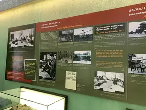 Đây là hình ảnh chụp một không gian trưng bày trong bảo tàng hoặc khu di tích lịch sử, với các tấm bảng thông tin lớn trên tường và một tủ kính trưng bày hiện vật. Các tấm bảng cung cấp thông tin và hình ảnh tư liệu về các sự kiện trong Chiến dịch Mùa Xuân 1975.。**Nội dung văn bản được ghi lại:**。**Bảng bên trái:**。*   Tiêu đề: "Ai chiếm được Tây Nguyên sẽ làm chủ Việt Nam và Đông Dương" (Đại tướng Võ Nguyên Giáp).。*   Nội dung: Trình bày về Hội nghị Bộ Chính trị mở rộng (18/12/1974 - 08/01/1975), nơi đã ra Nghị quyết lịch sử về kế hoạch giải phóng miền Nam trong hai năm 1975-1976 và nhấn mạnh việc giải phóng ngay trong năm 1975 nếu có thời cơ. Tây Nguyên được chọn làm nơi mở màn cuộc Tổng tiến công.。**Bảng ở giữa:**。*   Tiêu đề: "04/03 - 24/03/1975 GIẢI PHÓNG TÂY NGUYÊN".。*   Nội dung: Mô tả việc Bộ Chính trị cử Đại tướng Văn Tiến Dũng vào chiến trường. Các chú thích ảnh ghi lại các hoạt động: Đại tướng Văn Tiến Dũng bàn phương án hậu cần (tháng 02/1975); Đại tướng tại Sở Chỉ huy Mặt trận Tây Nguyên (tháng 03/1975); Bộ Tư lệnh Chiến dịch họp thông qua phương án đánh chiếm Buôn Ma Thuột (25/02/1975). Các hình ảnh khác cho thấy quân giải phóng tiến vào sân bay Hòa Bình, làm chủ Sở Chỉ huy Sư đoàn 23 tại Buôn Ma Thuột. Có hình ảnh tờ báo Quân đội Nhân dân số 4988 ngày 23/03/1975 với tiêu đề "TÂY NGUYÊN THẮNG LỚN".。**Bảng bên phải:**。*   Tiêu đề: "25/03/19[75] GIẢI PHÓNG H[UẾ]" và "GIẢI PHÓNG MIỀN NAM TRONG NĂM 1975".。*   Nội dung: Khi chiến dịch Tây Nguyên thắng lợi, Bộ Chính trị và Quân ủy Trung ương đã họp gấp tại Tổng Hành dinh (Nhà D67) để chuyển cuộc tiến công chiến lược thành Tổng tiến công chiến lược, giải phóng miền Nam trong năm 1975. Các hình ảnh cho thấy quân giải phóng tiến vào cửa Ngọ Môn (Thành nội Huế) và nhân dân thành phố Huế mít tinh mừng giải phóng. Một tấm ảnh khác ghi lại Hội nghị Bộ Chính trị mở rộng vào cuối tháng 03/1975.。**Cảnh vật và hoạt động:**。*   **Loại địa điểm:** Phòng trưng bày bảo tàng lịch sử.。*   **Hoạt động:** Trưng bày các tư liệu, hình ảnh và hiện vật về lịch sử quân sự Việt Nam, cụ thể là Chiến dịch Mùa Xuân 1975.。*   **Vật thể:** Các tấm pano thông tin, các bức ảnh tư liệu đen trắng về các cuộc họp, các trận đánh và hoạt động của quân dân. Bên dưới có một tủ kính trưng bày một bộ quân phục màu xanh được gấp lại.。（日本語訳）。これは博物館または史跡の展示スペースを撮影した写真で、壁には大きな情報パネルがあり、ガラスケースには遺物が展示されています。パネルは1975年春季攻勢作戦中の出来事に関する情報と資料写真を提供しています。**記録されているテキストの内容：**。**左側のパネル：**。*   タイトル：「テイグエンを制する者はベトナムとインドシナを制する」（ヴォー・グエン・ザップ大将）。*   内容：拡大政治局会議（1974年12月18日～1975年1月8日）について説明。この会議では、1975年から1976年の2年間で南部を解放する計画に関する歴史的な決議が採択され、好機があれば1975年中に解放することが強調された。テイグエンが総攻撃の開始地点として選ばれた。**中央のパネル：**。*   タイトル：「1975年3月4日～3月24日 テイグエン解放」。*   内容：政治局がヴァン・ティエン・ズン大将を戦場に派遣したことを記述。写真のキャプションには、ヴァン・ティエン・ズン大将が後方支援計画を協議する様子（1975年2月）、テイグエン戦線司令部にいる大将（1975年3月）、作戦司令部がバンメトート攻略計画を承認する会議（1975年2月25日）などの活動が記録されている。他の写真には、解放軍がホアビン空港に進軍し、バンメトートの第23師団司令部を制圧する様子が写っている。1975年3月23日付の人民軍新聞第4988号の写真があり、「テイグエン大勝利」という見出しがついている。**右側のパネル：**。*   タイトル：「19[75]年3月25日 フ[エ]解放」および「1975年中に南部を解放」。*   内容：テイグエン作戦が勝利すると、政治局と中央軍事委員会は総司令部（D67号棟）で緊急会議を開き、戦略的攻撃を総攻撃へと転換し、1975年中に南部を解放することを決定した。写真には、解放軍が午門（フエ王宮）に進軍する様子や、フエ市民が解放を祝って集会を開く様子が写っている。別の写真は1975年3月末の拡大政治局会議を記録している。**風景と活動：**。*   **場所の種類：**歴史博物館の展示室。*   **活動：**ベトナムの軍事史、具体的には1975年春季攻勢作戦に関する資料、写真、遺物の展示。*   **物体：**情報パネル、会議、戦闘、軍民の活動に関する白黒の資料写真。下には、折りたたまれた緑色の軍服一式を展示するガラスケースがある。