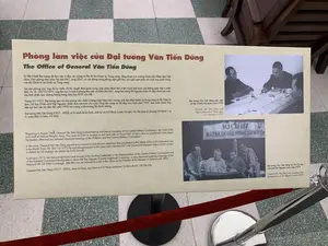 Đây là hình ảnh chụp một bảng thông tin được trưng bày trong một bảo tàng hoặc khu di tích lịch sử. Bảng thông tin này có tiêu đề chính bằng chữ màu nâu: "Phòng làm việc của Đại tướng Văn Tiến Dũng".。Nội dung văn bản tiếng Việt trên bảng được ghi lại đầy đủ như sau:。"Từ 08/1968 Đại tướng đã làm việc ở đây với cương vị Phó Bí thư Quân ủy Trung ương, Tổng tham mưu trưởng Quân đội Nhân dân Việt Nam. Căn phòng nằm phía Tây nhà D67, rộng 35m², có cửa thông sang phòng nghỉ giải lao, nơi nghỉ ngơi giữa các cuộc họp quan trọng của Bộ Chính trị và Quân uỷ Trung ương.。Tại căn phòng này, ông đã ký nhiều chỉ thị, quyết định quan trọng, góp phần đánh bại chiến tranh phá hoại của không quân Mỹ ở miền Bắc. Từ 18 - 30/12/1972, ông liên tục có mặt ở phòng làm việc và hầm nhà D67, cùng Đại tướng Võ Nguyên Giáp chỉ đạo đánh bại cuộc tập kích chiến lược đường không bằng máy bay B.52 của Mỹ.。Tháng 01/1975, Đại tướng tạm rời căn phòng vào chiến trường miền Nam trên cương vị là đại diện Quân uỷ Trung ương và Bộ Tổng Tư lệnh, chỉ huy Chiến dịch Tây Nguyên, chiến dịch mở màn cho cuộc Tổng tiến công và nổi dậy mùa Xuân năm 1975. Sau chiến tranh, ông tiếp tục làm việc tại đây đến năm 1992 với cương vị Bộ trưởng Bộ Quốc phòng.。Đại tướng Văn Tiến Dũng (1917 - 2002) có bí danh là Lê Hoài, sinh tại xã Cổ Nhuế, huyện Từ Liêm, Hà Nội (nay là phường Cổ Nhuế 1 và 2, quận Bắc Từ Liêm, Hà Nội)."。Trên bảng còn có hai bức ảnh tư liệu đen trắng kèm chú thích:。1. Bức ảnh trên cùng bên phải: Chụp cảnh một cuộc họp, với chú thích: "Đại tướng Văn Tiến Dũng làm việc với Bộ Tư lệnh Hải quân, năm 1970".。2. Bức ảnh dưới: Chụp một nhóm quân nhân đang xem xét tài liệu hoặc bản đồ. Phía sau họ có một tấm biển với dòng chữ "SỞ CHỈ HUY MẶT TRẬN GIẢI PHÓNG TÂY NGUYÊN". Chú thích cho ảnh này là: "Đại tướng Văn Tiến Dũng tại Sở Chỉ huy Mặt trận Tây Nguyên, mùa xuân 1975".。Bối cảnh xung quanh cho thấy sàn nhà lát gạch màu xám và trắng, cùng một cột chắn inox có dây nhung màu đỏ, xác nhận đây là một không gian trưng bày.。（日本語訳）。これは、博物館または史跡に展示されている案内板を撮影した写真です。この案内板には、茶色の文字で「ヴァン・ティエン・ズン大将の執務室」という主要なタイトルが付いています。案内板に記載されているベトナム語のテキストの全文は以下の通りです。「1968年8月から、大将は中央軍事委員会副書記、ベトナム人民軍総参謀長としてここで勤務しました。この部屋はD67棟の西側に位置し、広さは35平方メートルで、政治局と中央軍事委員会の重要な会議の合間に休憩する休憩室に通じるドアがあります。この部屋で、彼は多くの重要な指令や決定に署名し、北部における米空軍による破壊戦争の打破に貢献しました。1972年12月18日から30日まで、彼は執務室とD67棟の地下壕に常駐し、ヴォー・グエン・ザップ大将と共に、米国のB-52爆撃機による戦略的空爆の撃退を指揮しました。1975年1月、大将は中央軍事委員会および総司令部の代表として一時的にこの部屋を離れ、南部戦線へ赴き、1975年春の総攻撃と蜂起の幕開けとなったテイグエン作戦を指揮しました。戦後、彼は国防大臣として1992年までここで勤務を続けました。ヴァン・ティエン・ズン大将（1917年～2002年）は、レ・ホアイという偽名を持ち、ハノイ市トゥーリエム県コーニュエ社（現在のハノイ市バックトゥーリエム区コーニュエ1坊および2坊）で生まれました。」。案内板には、キャプション付きの2枚の白黒の資料写真もあります。1. 右上の写真：会議の様子を撮影したもので、「ヴァン・ティエン・ズン大将、海軍司令部と協議、1970年」というキャプションが付いています。2. 下の写真：軍人グループが資料または地図を調べている様子を撮影したもの。彼らの後ろには「テイグエン解放戦線司令部」と書かれた看板があります。この写真のキャプションは、「ヴァン・ティエン・ズン大将、テイグエン戦線司令部にて、1975年春」です。周囲の背景には、灰色と白のタイル張りの床と、赤いベルベットのロープが付いたステンレス製のポールパーテーションが見え、ここが展示スペースであることを示しています。