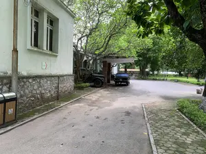 Khung cảnh chụp một con đường trải nhựa trong một khuôn viên có nhiều cây xanh. Bên trái là một tòa nhà cũ, tường sơn màu trắng ngà, phần chân tường được ốp bằng đá hộc. Trên tường tòa nhà có một tấm biển chỉ dẫn màu xanh lá cây ghi chữ 'HƯỚNG THAM QUAN' và 'KHU TỔ HỢP' với một mũi tên màu đỏ chỉ sang phải. Dưới một mái che, có hai chiếc ô tô kiểu cũ đang đỗ: một chiếc xe tải quân sự màu xanh đậm và một chiếc xe sedan màu đen. Con đường được trải nhựa, bên cạnh là vỉa hè lát gạch block hình lục giác. Có một thùng rác công cộng với thân ốp gỗ và nắp inox đặt trên vỉa hè. Khung cảnh vắng lặng, không có hoạt động nào của con người đang diễn ra.。（日本語訳）。緑豊かな敷地内にある舗装された道を撮影した風景。左側には古い建物があり、壁はアイボリーホワイトに塗られ、壁の根元部分は粗石で覆われている。建物の壁には、「HƯỚNG THAM QUAN」と「KHU TỔ HỢP」と書かれた緑色の案内板があり、右を指す赤い矢印が付いている。屋根の下には、2台の旧式の車が駐車している。1台は深緑色の軍用トラック、もう1台は黒色のセダンである。道路は舗装されており、その隣には六角形のブロックタイルで舗装された歩道がある。風景は静まり返っており、人の活動は見られない。