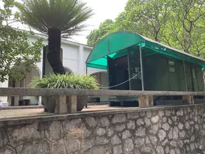 Trên thành toa xe lửa màu xanh có ghi chữ và số 'BT 58-10'. Bối cảnh là một khu trưng bày ngoài trời, có thể là trong khuôn viên của một bảo tàng hoặc di tích lịch sử. Hiện vật chính là một toa xe lửa cũ, được sơn màu xanh quân đội, đặt dưới một mái che bằng tấm nhựa màu xanh lá cây. Cửa toa xe đang mở nhưng được chắn lại bằng một sợi xích. Phía trước là một bức tường xây bằng đá và một chậu cây cảnh lớn trồng cây lan chi. Bên cạnh đó là một cây vạn tuế. Phía sau toa xe là một phần của một tòa nhà màu trắng với hành lang có mái vòm. Xung quanh có nhiều cây xanh tươi tốt.。（日本語訳）。緑色の鉄道車両の側面には、「BT 58-10」という文字と数字が書かれている。背景は屋外の展示エリアで、博物館または史跡の敷地内である可能性がある。車両のドアは開いているが、一本の鎖で塞がれている。手前には石で造られた壁と、オリヅルランが植えられた大きな観葉植物の鉢がある。その隣にはソテツの木がある。周囲には青々とした緑の木々がたくさんある。