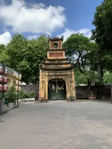 Bức ảnh chụp một cổng thành cổ kính, sơn màu vàng, mang đậm nét kiến trúc truyền thống. Cổng có một vọng lâu ở phía trên với mái cong nhiều tầng và các chi tiết trang trí. Cánh cửa chính bằng gỗ, sơn màu xanh lá cây và đang đóng. Xung quanh cổng là những bức tường gạch và nhiều cây xanh tươi tốt. Bầu trời trong xanh với những đám mây trắng. Bên trái cổng có một phần của một tòa nhà với biển hiệu ghi chữ '... & Cafe' và một cột treo nhiều đèn lồng trang trí. Một thùng rác màu xanh có dòng chữ 'HÃY BỎ RÁC VÀO THÙNG' cũng được nhìn thấy. Bên phải cổng có một chậu cây cảnh bonsai. Khung cảnh vắng lặng, không có hoạt động nào của con người đang diễn ra.。（日本語訳）。写真は、伝統的な建築様式が色濃く反映された、黄色に塗られた古風な城門を写している。門の上部には、幾層にも重なる反りのある屋根と装飾的な細部を持つ望楼がある。正面の扉は木製で、緑色に塗られており、閉まっている。門の周りにはレンガの壁と、青々と茂る多くの木々がある。空は青く澄み渡り、白い雲が浮かんでいる。門の左側には、「... & Cafe」と書かれた看板のある建物の一部と、多くの装飾的な提灯が吊るされた柱がある。「HÃY BỎ RÁC VÀO THÙNG」（ゴミはゴミ箱に入れてください）という文字が書かれた青いゴミ箱も見られる。門の右側には盆栽の鉢植えがある。辺りは静まり返っており、人の活動は見られない。