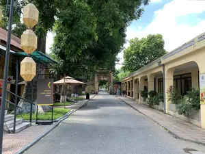 Bản ghi chữ viết: Một biển chỉ dẫn màu đen với chữ màu vàng ghi 'PHÒNG CHIẾU PHIM' và 'CINEMA', cùng với một mũi tên chỉ sang trái. Có các biển báo khác gần cổng thành và trên các tòa nhà nhưng chữ viết không thể đọc rõ.。Mô tả cảnh vật và hoạt động: Bức ảnh chụp một con đường trải nhựa yên tĩnh bên trong một khu di tích lịch sử. Hai bên đường là những hàng cây cổ thụ xanh tốt. Bên phải là một dãy nhà dài một tầng sơn màu vàng với hàng hiên có mái vòm, có thể là khu dịch vụ hoặc quán cà phê cho khách tham quan. Bên trái có một cột đèn treo những chiếc đèn lồng trang trí màu vàng với hoa văn màu đỏ. Ở cuối con đường là một chiếc cổng thành cổ kính xây bằng gạch. Khung cảnh khá vắng vẻ, chỉ có hai người đang đi bộ ở phía xa, cho thấy hoạt động tham quan đang diễn ra.。（日本語訳）。文字の記録：「PHÒNG CHIẾU PHIM」と「CINEMA」と書かれた黄色の文字と、左を指す矢印がある黒い案内板。城門の近くや建物の上にも他の看板があるが、文字ははっきりと読み取れない。風景と活動の説明：写真は、歴史的遺跡地区内の静かなアスファルトの道を撮影したものである。道の両側には、青々とした古い木々の並木がある。右側には、アーチ型の屋根を持つベランダ付きの黄色く塗られた長い平屋の建物があり、観光客向けのサービスエリアまたはカフェである可能性がある。左側には、赤い模様の入った黄色い装飾的な提灯が吊るされた街灯がある。道の突き当たりには、レンガで造られた古風な城門がある。風景はかなり閑散としており、遠くに歩いている人が2人だけいることから、観光活動が行われていることがわかる。