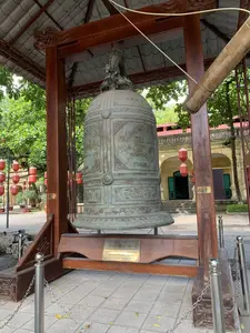 Bức ảnh chụp một quả chuông đồng lớn (Đại hồng chung) được đặt trong một nhà chuông tại một khu di tích lịch sử, có thể là Hoàng thành Thăng Long. Quả chuông được treo trên một bộ giá đỡ bằng gỗ lớn, có mái ngói che. Các đầu xà của giá đỡ được chạm khắc hình đầu rồng. Bên cạnh chuông có một khúc gỗ lớn dùng để đánh chuông. Quả chuông có màu đồng cổ, trên thân có khắc chữ và hoa văn. Phía trước, trên giá đỡ có gắn một tấm biển kim loại màu vàng ghi thông tin về quả chuông. Xung quanh chuông có hàng rào bằng cọc inox và dây xích. Phía sau là sân và một tòa nhà cổ có tường vàng, cửa chớp xanh. Nhiều đèn lồng đỏ được treo xung quanh, tạo nên không khí trang nghiêm. Không có hoạt động của người nào được ghi lại.。Nội dung chữ viết trên tấm biển kim loại:。ĐẠI HỒNG CHUNG。Chất liệu: Đúc bằng đồng đỏ. Cao: 3050mm - Trọng lượng: 3470kg。HIỆN VẬT ĐẶC BIỆT CỦA CỘNG ĐỒNG LÀNG NGHỀ ĐÚC ĐỒNG THỦ CÔNG MỸ NGHỆ HÀ NỘI。CÁC CƠ QUAN BAN NGÀNH, CÁC DOANH NGHIỆP, ĐÔNG ĐẢO TRONG VÀ NGOÀI NƯỚC ĐÓNG GÓP。CUNG TIẾN LÊN ĐIỆN KÍNH THIÊN - HOÀNG THÀNH THĂNG LONG - HÀ NỘI。CÔNG TRÌNH KHỞI CÔNG THÁNG 8 - 2010. HOÀN THÀNH THÁNG 9 - 2010。Trên thân chuông có khắc chữ Hán '昇龍大鐘' (Thăng Long Đại Chung).。（日本語訳）。写真は、歴史的遺跡（おそらくタンロン皇城）にある鐘楼に置かれた大きな銅の鐘（大洪鐘）を写している。鐘は、瓦屋根で覆われた大きな木製の架台に吊るされている。架台の梁の端は、龍の頭の形に彫刻されている。鐘の隣には、鐘を打つために使われる大きな丸太がある。鐘は古銅色をしており、胴体には文字と文様が刻まれている。前方の架台には、鐘に関する情報が記された金色の金属プレートが取り付けられている。鐘の周りには、ステンレスの杭と鎖でできた柵がある。後ろには中庭と、黄色い壁に緑色の鎧戸がある古い建物がある。多くの赤い提灯が周りに吊るされており、荘厳な雰囲気を醸し出している。人の活動は記録されていない。金属プレートに書かれた文字の内容：。大洪鐘。材質：赤銅で鋳造。高さ：3050mm - 重量：3470kg。ハノイ手工芸銅鋳造職人村共同体の特別な現物。各機関・部門、各企業、国内外の多数の方々が寄進。ハノイ - タンロン皇城 - 敬天殿へ奉納。工事着工 2010年8月。完成 2010年9月。鐘の胴体には漢字「昇龍大鐘」（タンロン・ダイ・チュン）が刻まれている。