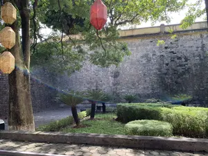 Bức ảnh chụp một khung cảnh ngoài trời yên tĩnh tại một khu di tích lịch sử, có khả năng là Hoàng thành Thăng Long. Nổi bật ở hậu cảnh là một bức tường thành cổ, xây bằng gạch đã nhuốm màu thời gian. Phía trước bức tường là một khu vườn nhỏ được chăm sóc với các bụi cây được cắt tỉa gọn gàng, một vài cây vạn tuế và thảm cỏ xanh. Ở bên trái có một cây cổ thụ lớn, trên cành cây có treo những chiếc đèn lồng trang trí mang phong cách Á Đông, bao gồm một chiếc màu đỏ và một chuỗi đèn lồng màu vàng có hoa văn. Ánh nắng mặt trời chiếu xuyên qua tán lá, tạo ra một vệt sáng giống như cầu vồng. Lối đi phía trước được lát bằng gạch block. Không có hoạt động của con người được ghi lại, khung cảnh mang lại cảm giác thanh bình.。（日本語訳）。写真は、おそらくタンロン皇城と思われる史跡の、静かな屋外の風景を撮影したものです。背景には、時を経て色づいたレンガで造られた古い城壁が際立っています。城壁の前には、きれいに刈り込まれた低木、数本のソテツ、そして緑の芝生がある、手入れの行き届いた小さな庭があります。左側には大きな古木があり、その枝には東アジア風の装飾的な提灯が吊るされています。それには、赤い提灯が一つと、模様の入った黄色い提灯の連なりが含まれます。太陽の光が木の葉を突き抜けて差し込み、虹のような光の筋を作り出しています。手前の通路はブロックレンガで舗装されています。人の活動は見られず、その光景は平穏な感覚をもたらします。