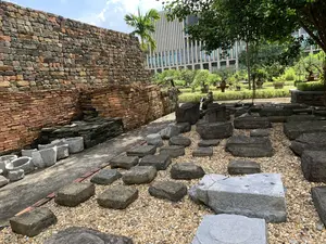 Bức ảnh chụp một khu di tích khảo cổ học ngoài trời vào ban ngày. Bên trái là một bức tường gạch cổ, có thể là một phần của một công trình kiến trúc xưa đã được khai quật. Bức tường được xây bằng các viên gạch có màu sắc đa dạng. Dọc theo chân tường là một hàng các máng hoặc chậu bằng đá. Phần trung tâm và bên phải của bức ảnh là một khu vực rải sỏi, trên đó đặt nhiều khối đá lớn, vuông vức, có thể là đá tảng kê chân cột hoặc đá lát nền của một công trình cổ. Ở hậu cảnh, phía sau khu di tích là một khu vườn được chăm sóc cẩn thận với cây xanh, cỏ và nhiều chậu cây cảnh. Xa hơn nữa là một tòa nhà hiện đại, cao tầng với mặt tiền bằng kính, tạo ra sự tương phản rõ rệt giữa quá khứ và hiện tại. Không có hoạt động nào của con người được ghi lại.。（日本語訳）。日中の屋外にある考古学遺跡を撮影した写真です。左側には古いレンガの壁があり、発掘された古代の建築物の一部である可能性があります。壁は様々な色のレンガで造られています。壁の麓に沿って、石製の樋または鉢が並んでいます。写真の中央と右側は砂利が敷かれたエリアで、その上には多くの大きな四角い石のブロックが置かれています。これらは柱の礎石か、古代の建物の敷石である可能性があります。背景、遺跡の後ろには、緑の木々、草、そして多くの鉢植えの観葉植物がある、手入れの行き届いた庭園があります。さらにその奥には、ガラス張りのファサードを持つ近代的な高層ビルがあり、過去と現在の明確な対比を生み出しています。人間の活動は記録されていません。