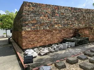 Bức ảnh chụp một khu vực trưng bày khảo cổ ngoài trời, có khả năng là một phần của khu di tích Hoàng thành Thăng Long. Trung tâm của bức ảnh là một đoạn tường thành cổ được xây bằng gạch và đá. Bức tường có hai phần khác nhau: phần thân dưới được xây bằng những viên gạch nhỏ màu nâu đỏ, trong khi phần trên được xây bằng những viên gạch đá lớn hơn với nhiều màu sắc như xám, đen và nâu. Phía trước bức tường, trên một khoảng sân xi măng, trưng bày nhiều hiện vật khảo cổ bằng đá. Các hiện vật này bao gồm các chân tảng cột đá có hình tròn và lỗ ở giữa, các loại cối hoặc chậu đá, và các khối đá hình chữ nhật. Bối cảnh là ngoài trời vào một ngày nắng đẹp với bầu trời xanh và cây cối xanh tươi ở phía sau. Có một bảng thông tin màu đen đặt trên giá đỡ nhưng không thể đọc được nội dung. Một chiếc đèn pha màu đen cũng được đặt ở tiền cảnh để chiếu sáng các hiện vật. Không có hoạt động nào của con người diễn ra, đây là một cảnh quan tĩnh tại một địa điểm lịch sử.。（日本語訳）。写真は屋外の考古学展示エリアを撮影したもので、おそらくタンロン皇城遺跡の一部である。写真の中央には、レンガと石で造られた古い城壁の一部がある。城壁は二つの異なる部分から成る。下部は赤褐色の小さなレンガで造られている一方、上部は灰色、黒、茶色など様々な色のより大きな石レンガで造られている。城壁の前、セメントの中庭には、多くの石造りの考古学的な遺物が展示されている。これらの遺物には、中央に穴の開いた円形の礎石、様々な種類の石臼や石鉢、そして長方形の石のブロックが含まれる。人の活動は見られず、これは史跡における静的な景観である。