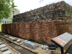 Bức ảnh chụp một khu trưng bày khảo cổ học ngoài trời, có khả năng là tại khu di tích Hoàng thành Thăng Long. Cảnh vật cho thấy một mặt cắt của một bức tường cổ đã được khai quật. Bức tường này có hai phần rõ rệt: phần trên được xây bằng gạch khối lớn, sẫm màu, trong khi phần dưới được tạo thành từ rất nhiều lớp ngói đất nung màu nâu đỏ xếp chồng lên nhau. Phía trước bức tường, trên nền sỏi đá, là các hiện vật bằng đá, có thể là các bộ phận kiến trúc cổ. Ở phía bên phải, có một bảng thông tin với tiêu đề 'VẬT LIỆU NGÓI CÁC THỜI KỲ' và dòng chữ tiếng Anh 'ROOF TILES IN DIFFERENT PERIODS'. Bảng này dường như giải thích về các loại vật liệu xây dựng được tìm thấy, tương ứng với các giai đoạn lịch sử khác nhau. Phía sau khu di tích là cây xanh, một cây cọ và một tòa nhà hiện đại, tạo ra sự tương phản giữa cổ xưa và đương đại. Không có hoạt động nào của con người được ghi lại; đây là một cảnh trưng bày tĩnh.。（日本語訳）。この写真は、屋外の考古学展示エリアを写したもので、おそらくタンロン皇城遺跡にあるものだと思われます。風景は、発掘された古い壁の断面を示しています。この壁には明確に二つの部分があります。上部は大きな暗色のレンガで造られ、下部は赤褐色のテラコッタ瓦が何層にも積み重なってできています。壁の前、砂利の地面の上には、石の遺物があり、おそらくは古い建築の部材でしょう。右側には、「VẬT LIỆU NGÓI CÁC THỜI KỲ」という見出しと、「ROOF TILES IN DIFFERENT PERIODS」という英語の文字が書かれた案内板があります。この案内板は、発見された建材の種類について、異なる歴史的時代に対応させて説明しているようです。遺跡の後ろには緑の木々、ヤシの木、そして現代的な建物があり、古代と現代の対比を生み出しています。人の活動は記録されていません。これは静的な展示風景です。