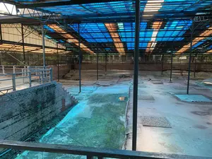 Đây là một địa điểm khảo cổ học rộng lớn, được che chắn bởi một kết cấu mái nhà lớn làm bằng khung kim loại và các tấm lợp mờ nhiều màu sắc, chủ yếu là màu xanh dương và màu hổ phách. Bên dưới mái che là khu vực khai quật với nền đất không bằng phẳng, cho thấy các lớp di tích khác nhau. Có thể nhìn thấy các bức tường gạch cũ, một số đã bị phong hóa, và một cấu trúc bậc thang bằng gạch ở phía bên trái. Nhiều phần của khu vực khai quật được phủ bằng các tấm bạt lớn, có thể để bảo vệ các hiện vật khỏi tác động của môi trường. Không có người trong ảnh và không có hoạt động nào đang diễn ra, cho thấy địa điểm này đang trong quá trình bảo tồn hoặc tạm nghỉ. Ở phía trước là một lan can kim loại, gợi ý rằng đây là một lối đi để quan sát khu vực khai quật từ trên cao. Không có văn bản hay chữ viết nào có thể nhận dạng được.。（日本語訳）。これは広大な考古学遺跡であり、金属フレームと、主に青色と琥珀色の多色半透明の屋根パネルで作られた大きな屋根構造で覆われています。屋根の下には、地面が不均一な発掘エリアがあり、さまざまな時代の遺跡の層が見られます。古いレンガの壁が見え、その一部は風化しており、左側にはレンガ造りの階段状の構造物があります。発掘エリアの多くの部分は大きな防水シートで覆われており、おそらく遺物を環境の影響から保護するためでしょう。写真には人がおらず、活動も行われていないことから、この場所が保存作業中か、一時的に休止していることを示唆しています。手前には金属製の手すりがあり、これが発掘エリアを上から観察するための通路であることを示唆しています。識別できるテキストや文字はありません。