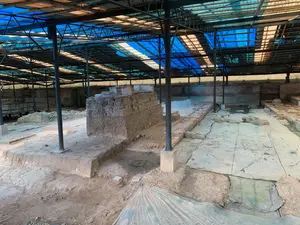 Đây là hình ảnh chụp một khu khai quật khảo cổ học. Toàn bộ khu vực được che bởi một mái che lớn có kết cấu khung thép và lợp bằng các tấm nhựa màu xanh và trong suốt. Bên dưới là hiện trường khai quật, để lộ ra các nền móng và tường gạch cổ đang trong quá trình nghiên cứu. Có một khối đất và gạch lớn ở trung tâm cho thấy các lớp địa tầng khác nhau. Một số khu vực được phủ bạt để bảo vệ di vật. Các cột thép được dựng lên để chống đỡ mái che. Không có hoạt động rõ ràng nào đang diễn ra, và không có văn bản hay chữ viết nào có thể nhìn thấy.。（日本語訳）。これは考古学の発掘現場を撮影した写真です。全域が、鉄骨構造で、青色と透明のプラスチック板で覆われた大きな屋根で覆われています。その下には発掘現場があり、調査中の古代の基礎やレンガの壁が露出しています。中央には、異なる地層を示す大きな土とレンガの塊があります。いくつかのエリアは、遺物を保護するために防水シートで覆われています。屋根を支えるために鉄骨の柱が立てられています。