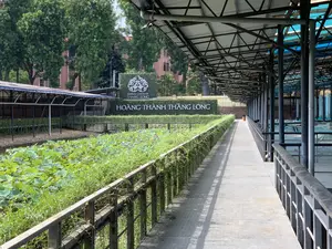 Trên một tấm biển lớn được làm bằng cây xanh có dòng chữ 'IMPERIAL CITADEL OF THANG LONG' và 'HOÀNG THÀNH THĂNG LONG'. Bối cảnh là một khu vực bên trong Hoàng Thành Thăng Long. Có một lối đi dài có mái che bằng khung kim loại. Bên trái lối đi là một con mương hoặc hồ nhỏ trồng đầy sen, có thể thấy nhiều lá sen xanh và một vài bông hoa sen màu hồng. Bên phải lối đi là một khu vực có mái che, trông giống như một địa điểm khai quật khảo cổ học đang được bảo vệ. Khung cảnh yên tĩnh, không có người. Ở phía sau là những hàng cây xanh và một tòa nhà.。（日本語訳）。緑の植物で作られた大きな看板に、「IMPERIAL CITADEL OF THANG LONG」と「HOÀNG THÀNH THĂNG LONG」という文字があります。背景はタンロン皇城内のエリアです。金属フレームで屋根が覆われた長い通路があります。通路の左側には、蓮が植えられた堀または小さな池があり、たくさんの緑の蓮の葉といくつかのピンクの蓮の花が見えます。通路の右側には屋根付きのエリアがあり、保護されている考古学の発掘現場のように見えます。景色は静かで、人はいません。後方には緑の木々の列と建物があります。