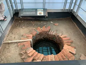 Đây là hình ảnh chụp một khu di tích khảo cổ, nhìn từ trên một sàn quan sát bằng kim loại xuống dưới. Đối tượng chính là một giếng nước cổ được khai quật. Cạnh khu vực giếng có một bảng thông tin với nội dung được ghi lại như sau:。Nội dung trên bảng thông tin:。Tiếng Việt: "GIẾNG NƯỚC THỜI TRẦN THẾ KỶ XIII - XIV. Giếng nước sâu 2,4m, được xây bằng gạch với kỹ thuật xếp gạch hình 'xương cá'. Khi khai quật, các nhà khảo cổ đã tìm thấy trong lòng giếng nhiều đồ gốm và vật liệu kiến trúc thời Trần. Đây là chiếc giếng gạch có kỹ thuật xây dựng độc đáo, duy nhất kiểu này trong khu di tích khảo cổ học 18 Hoàng Diệu."。Tiếng Anh: "WATER WELL OF THE TRAN DYNASTY (13TH - 14TH CENTURIES). The well is 2.4 meters deep, built of bricks in herringbone (fishbone) pattern. During excavations, archaeologists have found in the loop of the well a wide range of pottery and constructional materials that date back to the Tran Dynasty. In terms of construction technique, this is a unique and original brick well hitherto found in the Archaeological Site of 18 Hoang Dieu."。Mô tả cảnh vật và hoạt động:。- Địa điểm: Một khu khai quật khảo cổ, trưng bày một giếng nước cổ thời Trần.。- Hoạt động: Một du khách (chỉ thấy phần chân đi dép) đang đứng trên sàn kim loại để quan sát di tích giếng cổ bên dưới.。- Vật thể: Giếng cổ được xây bằng gạch nung màu đỏ theo kỹ thuật xếp lớp hình xương cá, nằm trong một hố đất. Miệng giếng không còn nguyên vẹn. Bên trong giếng có nước, phản chiếu khung lưới của sàn quan sát phía trên. Xung quanh giếng là nền đất của khu khai quật với vài mảnh gạch vỡ. Có một ống dẫn màu sáng (giống ống tre hoặc nhựa) chạy dọc theo mép hố.。（日本語訳）。これは考古学遺跡を撮影した写真で、金属製の展望台から下を見下ろしています。主な被写体は、発掘された古い井戸です。井戸のエリアの隣には、以下のように内容が記録された案内板があります。案内板の内容：。ベトナム語：「13～14世紀の陳朝時代の井戸。深さ2.4mの井戸で、「魚の骨」の形にレンガを積む技術で建てられている。発掘時、考古学者たちは井戸の中から陳朝時代の多くの陶器や建築資材を発見した。これは、ホアンジエウ18番地考古学遺跡地区において、この種の唯一の独創的な建設技術を持つレンガ造りの井戸である。」。英語：「陳朝時代（13～14世紀）の井戸。井戸は深さ2.4メートルで、ヘリンボーン（魚の骨）模様のレンガで造られている。発掘調査中、考古学者たちは井戸の内部で、陳朝時代に遡る多種多様な陶器や建築資材を発見した。建設技術の観点から、これはホアンジエウ18番地考古学遺跡でこれまでに発見された中で、ユニークで独創的なレンガ造りの井戸である。」。風景と活動の説明：。- 場所：陳朝時代の古い井戸を展示している考古学発掘現場。- 物体：古い井戸は、魚の骨の形に積む技術で赤レンガで建てられ、土の穴の中にある。井戸の口は完全な状態ではない。井戸の中には水があり、上にある展望台の格子状のフレームを映している。井戸の周りは発掘現場の地面で、いくつかの割れたレンガの破片がある。穴の縁に沿って、明るい色の導管（竹の管またはプラスチックの管に似ている）が走っている。